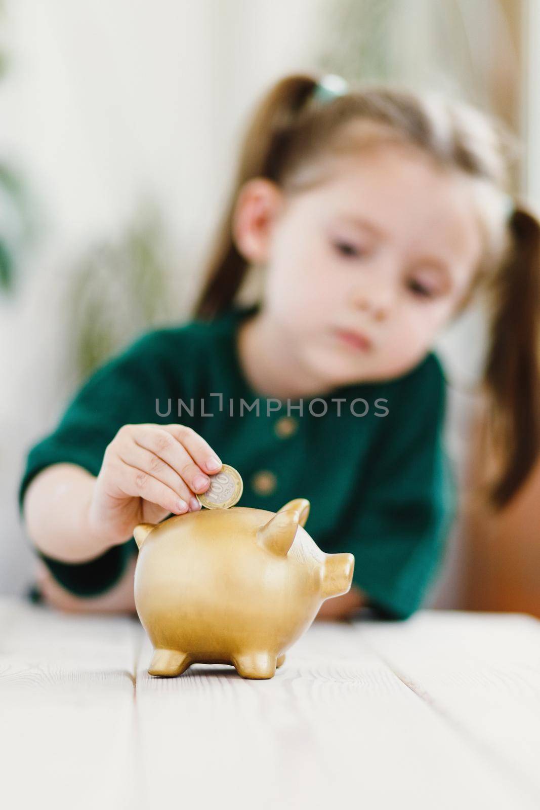 Close up view on child hand, she putting a coin in gold color piggy bank. Vertical. Selective soft focus