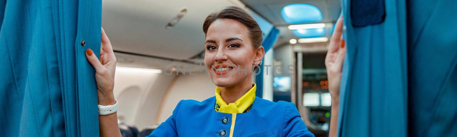 Cheerful flight attendant in air hostess uniform looking at camera and smiling while holding drapes in aircraft passenger salon