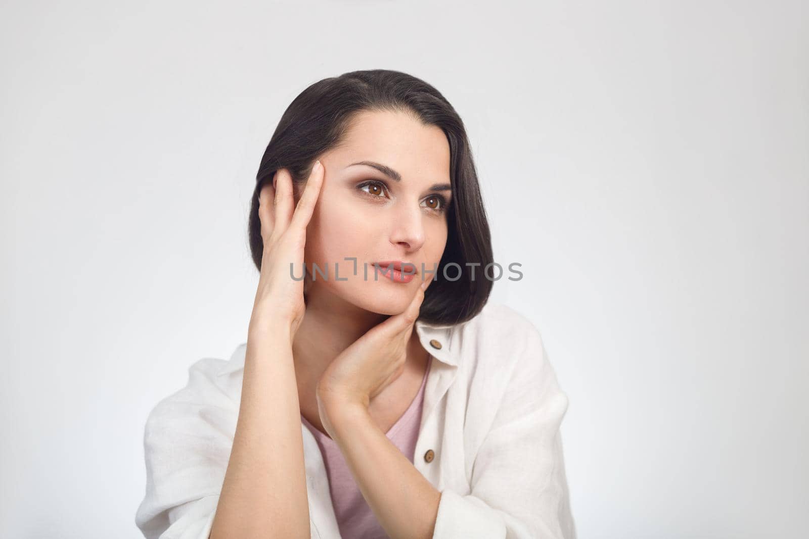 High-key headshot portrait of beautiful brunette female model with hands near face on white background.