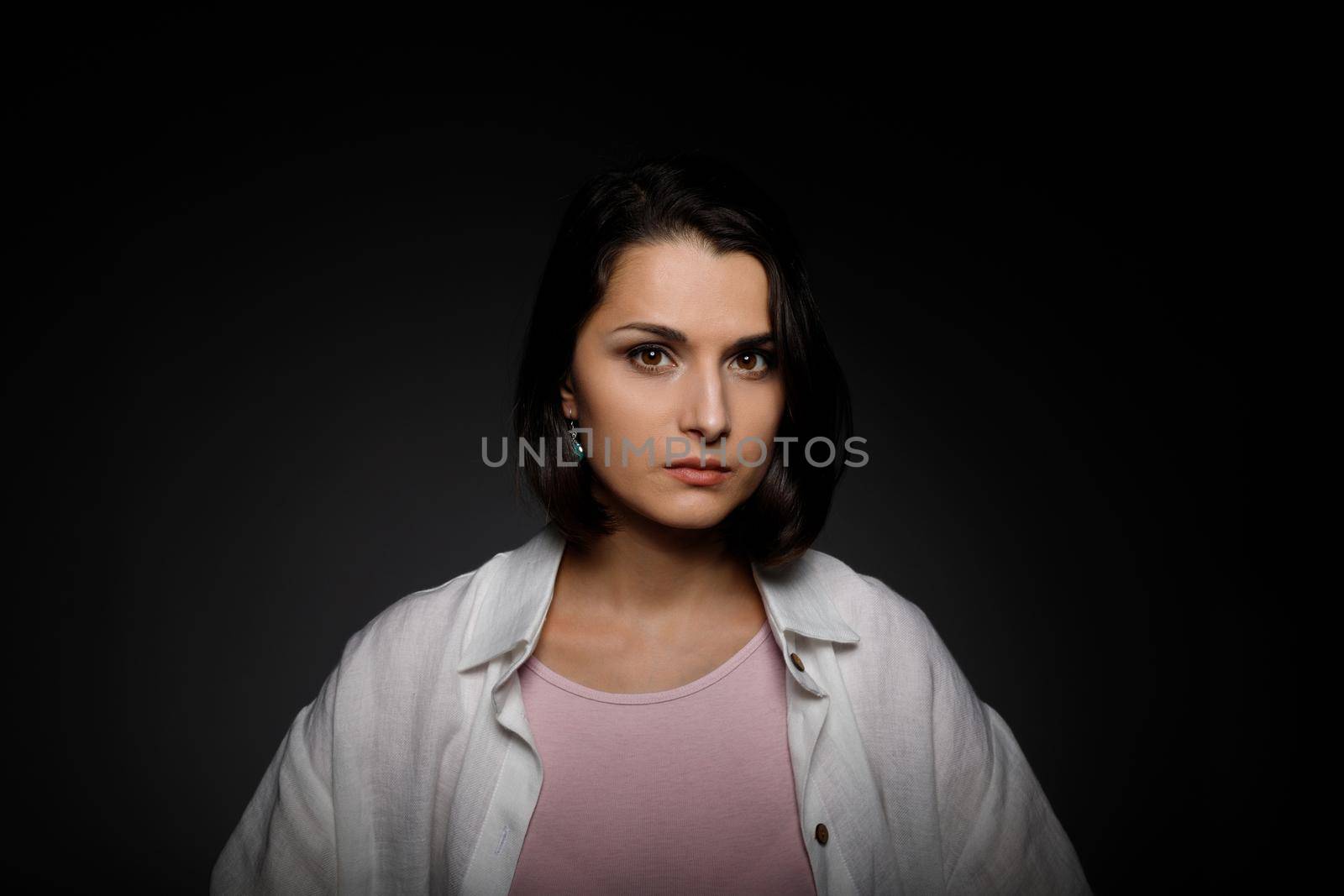 Low key portrait of serious young brunette multiethnic woman in pink tank and white shirt looking at camera.