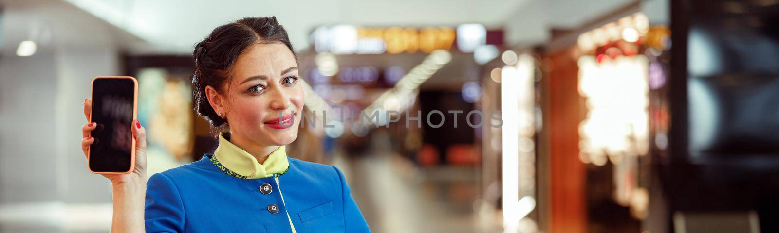 Cheerful woman flight attendant in aviation air hostess uniform looking at camera and smiling while holding smartphone