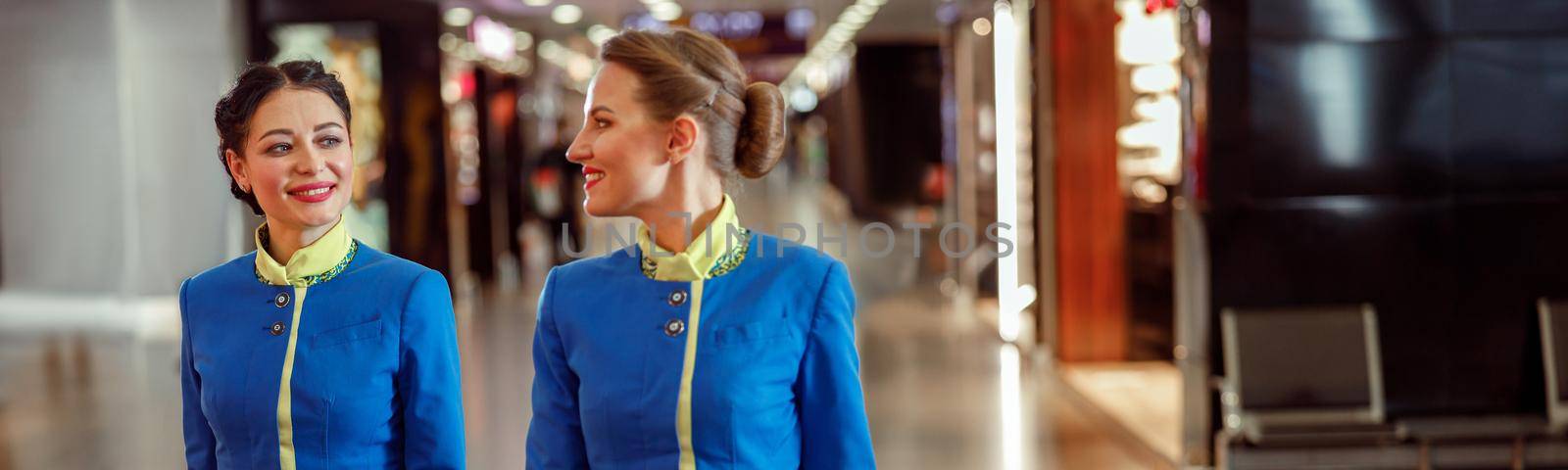 Two cheerful women stewardesses in air hostess uniform walking down airport terminal