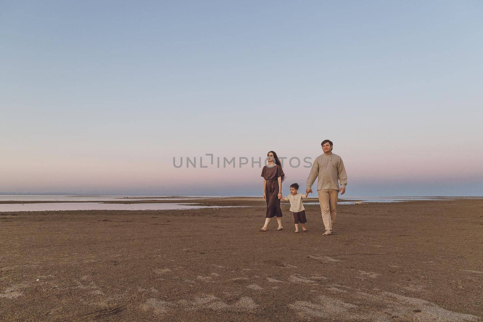 A young family with a little son in linen clothes are quietly walking along the evening beach. Copy space.