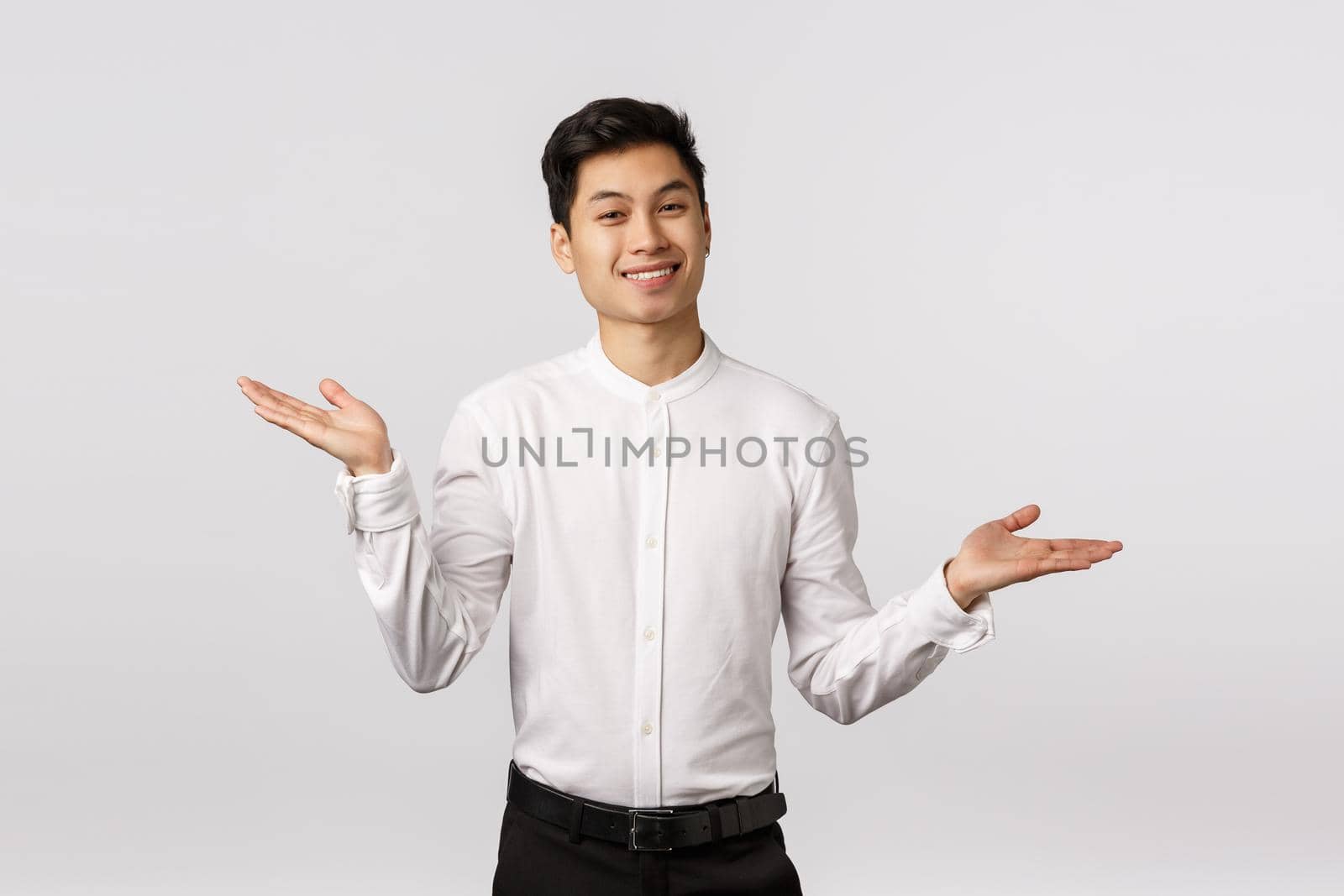 Unbothered and careless happy successful young male employee in white shirt, pants, spread hands sideways and shrugging with relaxed, relieved expression, smiling, dont know and dont care.