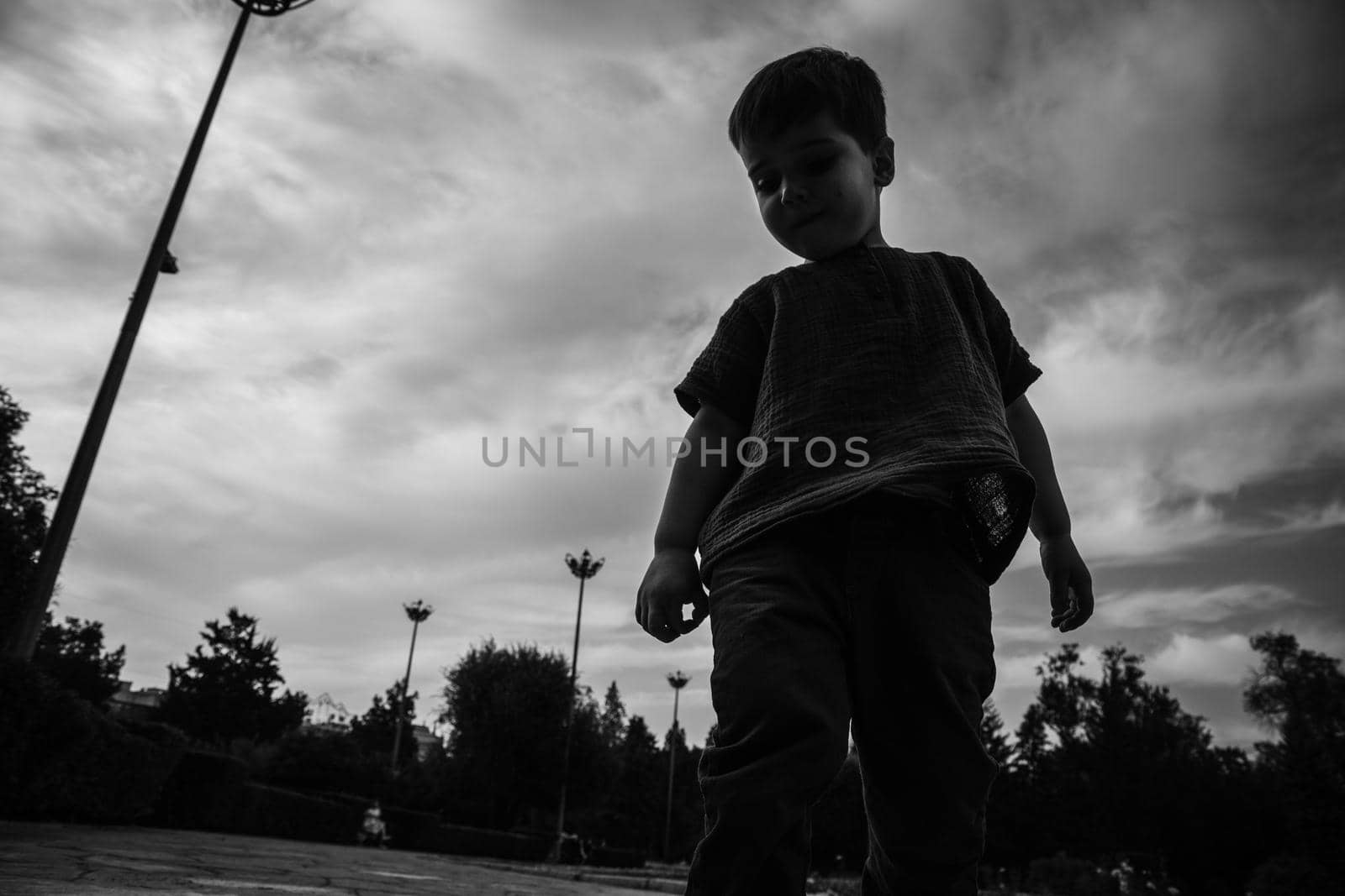 Black and white portrait of 3 year old boy in the park playing alone against the backdrop of a dramatic sky. BW lifestyle street photo.