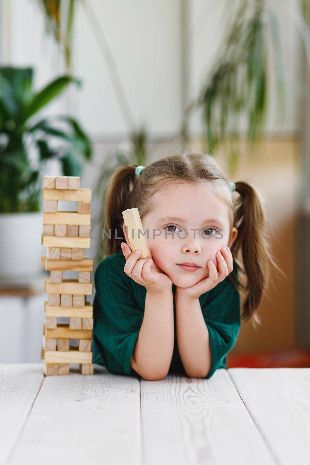 Portrait of beautiful sweet little girl in green dress with ponytails on her head holding a wooden block and wooden tower standing on a table.