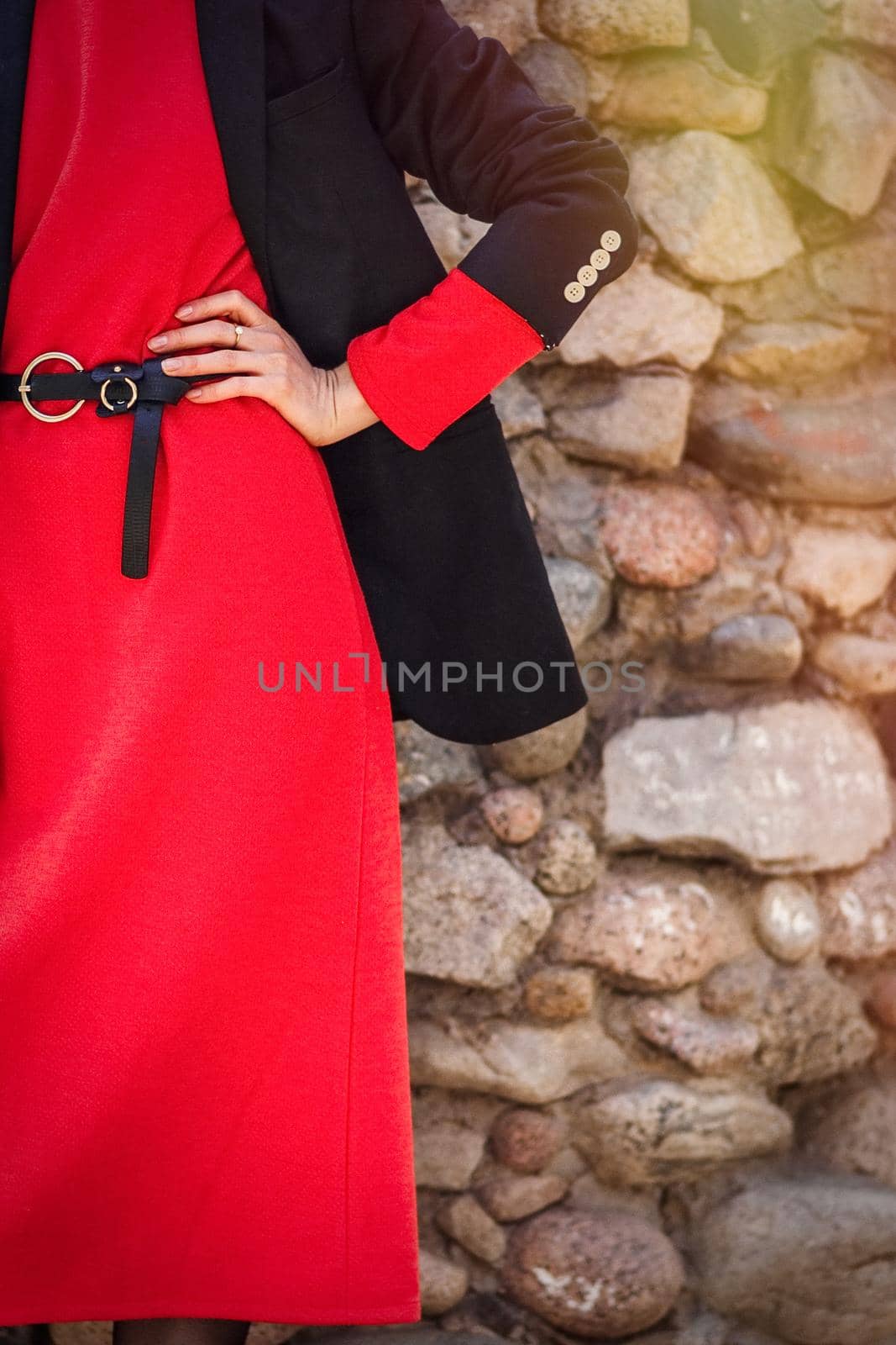 Close-up of a red dress, a black belt and a jacket on a woman against a background of a stone wall outdoor. Autumn fashion.
