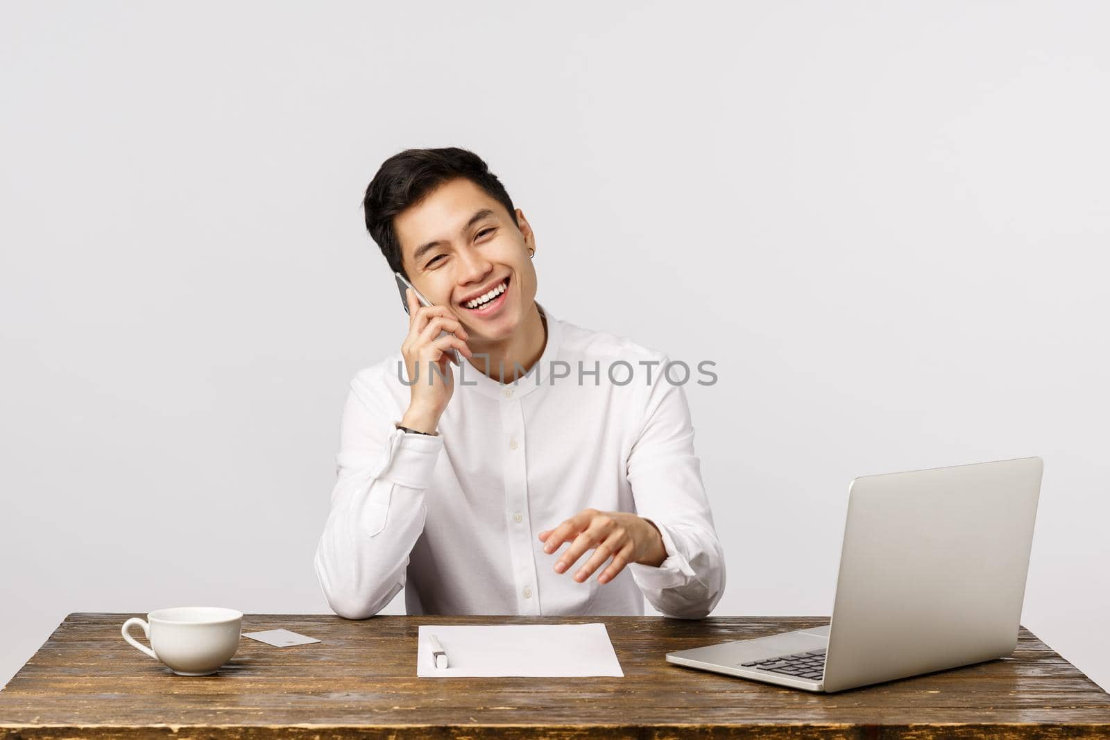 Carefree, smiling happy young asian businessman in office, sitting with laptop, documents, calling friend, talking and laughing as holding smartphone near ear, white background. Copy space