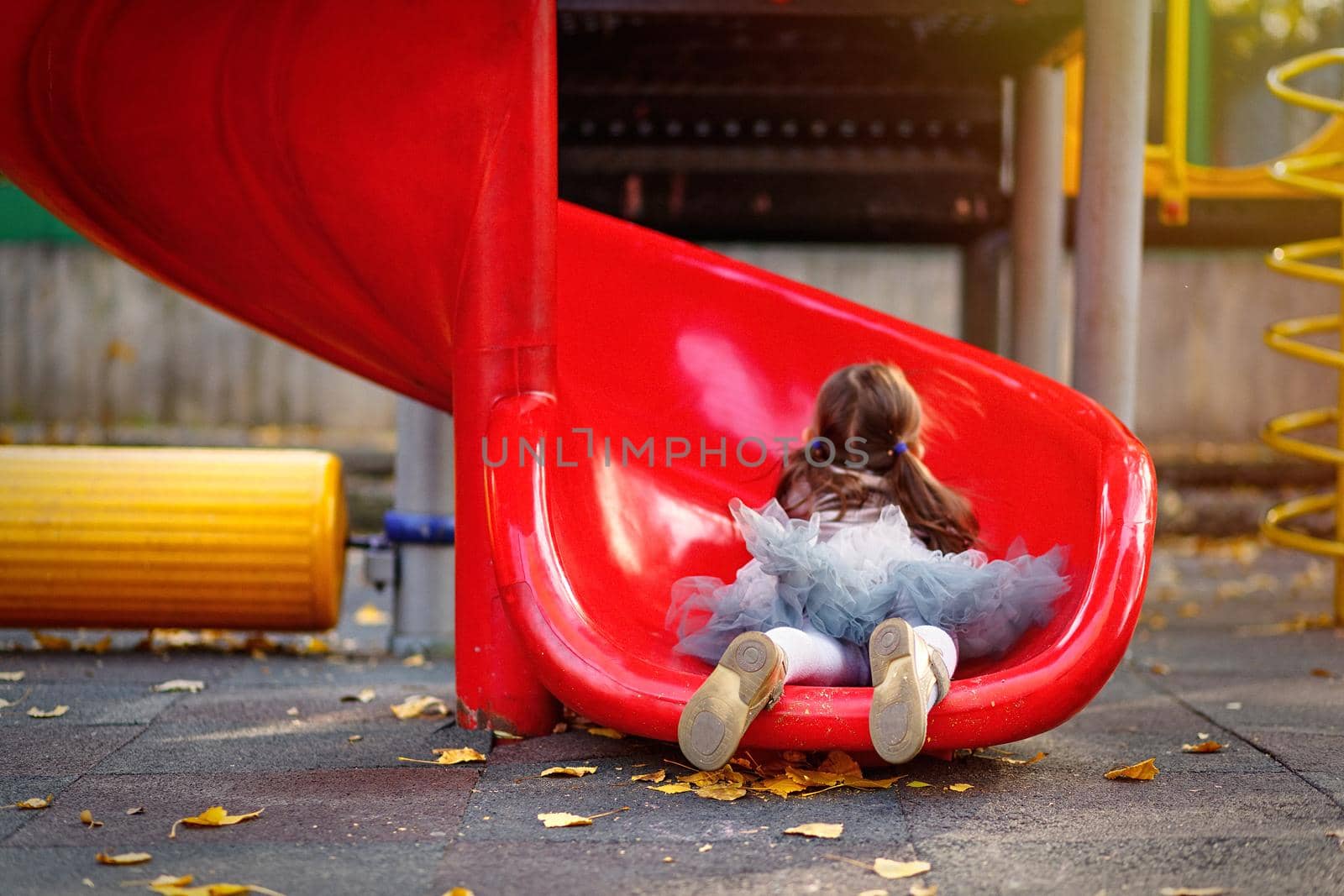 A cute girl in skirt tutu is riding a playground slide. Lifestyle outdoor portrait of a child. Motion blur and selective soft focus.