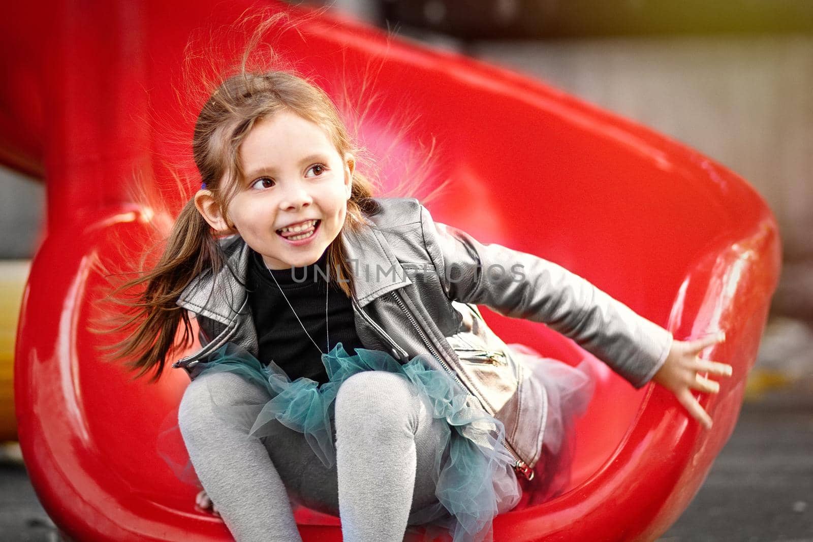 A cheerful girl is riding a slide in the park playground and laughing. Emotional outdoor portrait of a child. Motion blur and selective soft focus.