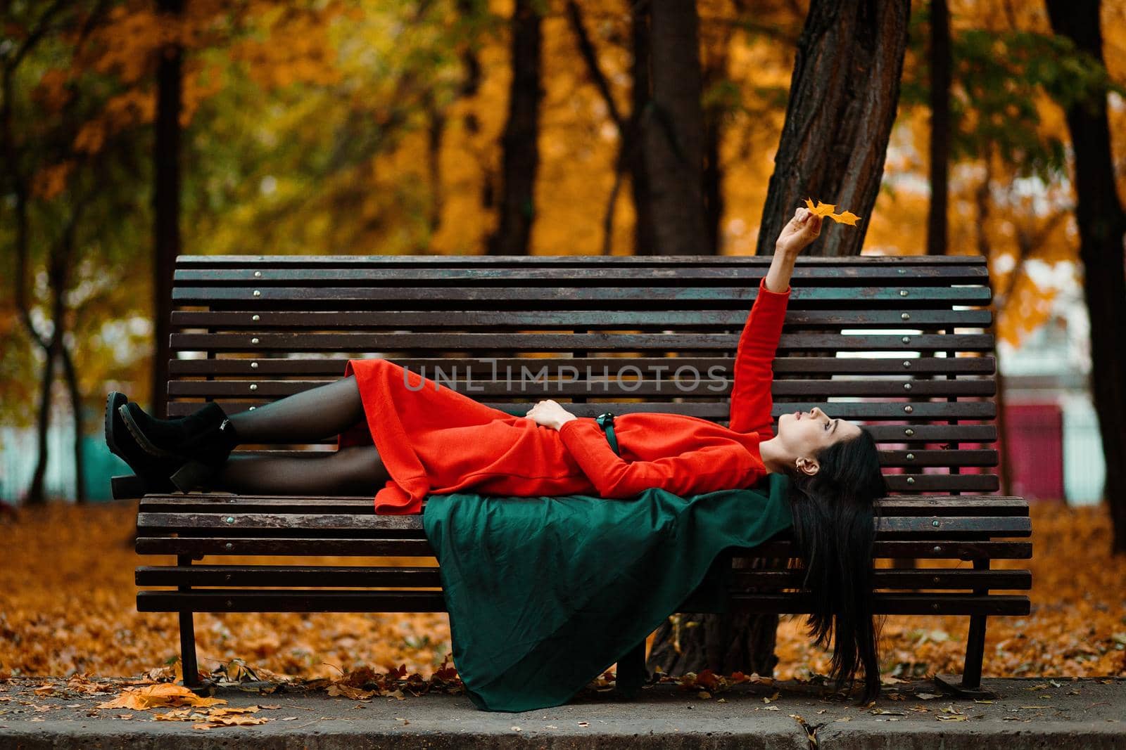 A young woman in an orange dress and a green coat lies on a bench in an autumn park and enjoys the autumn mood.