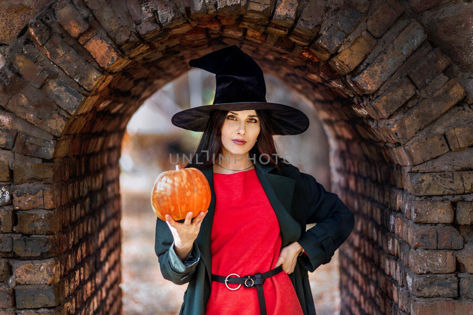 Beautiful young woman portrait in witch hat, holding a orange pumpkin. Halloween mood.