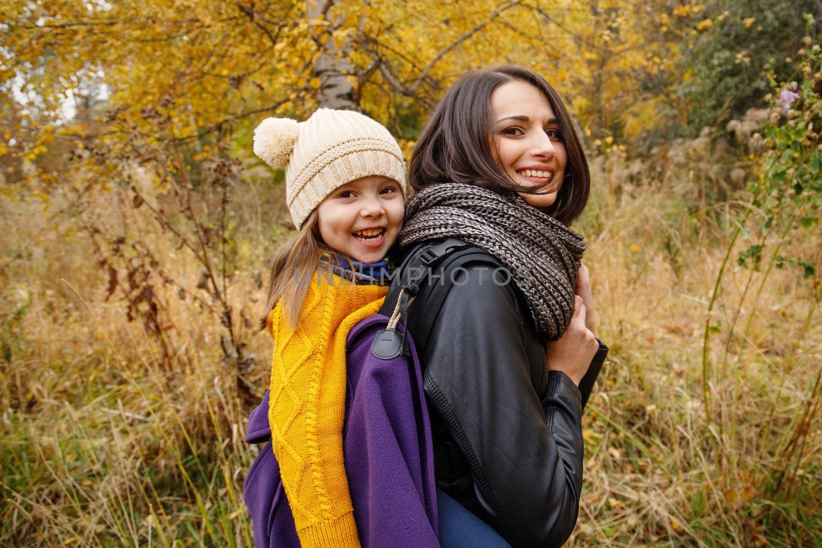 Happy young mother with her kid girl on back in ergonomic baby carrier in autumn nature. Babywearing concept.