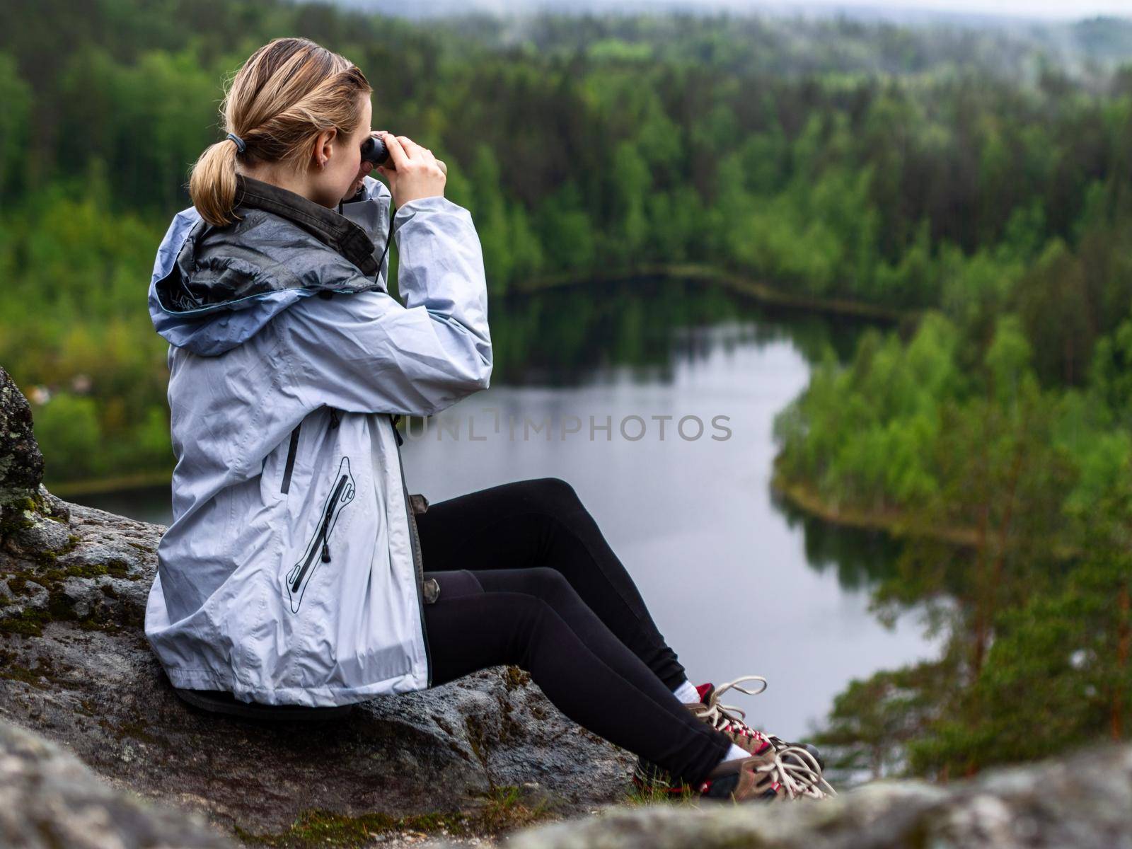 woman looks through binoculars. tourist girl on a rock in a forest near a lake