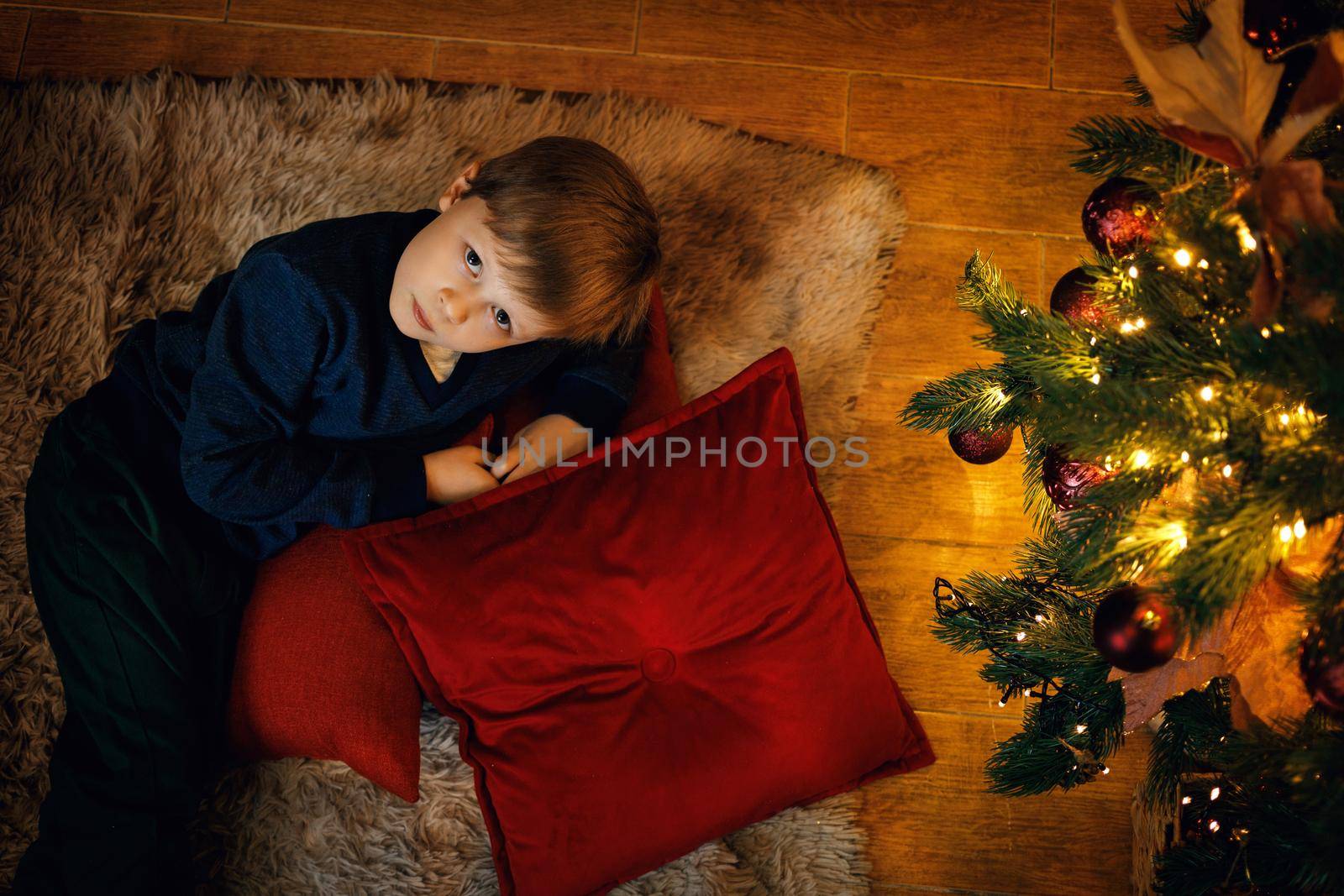 A blond boy 5-7 years old lies on the carpet near the New Year tree, looking at the camera. by Rom4ek