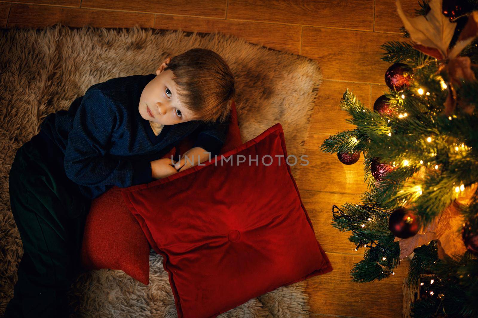 A blond boy 5-7 years old lies on the carpet near the New Year tree, looking at the camera. Top view. Selective soft focus, film grain effect