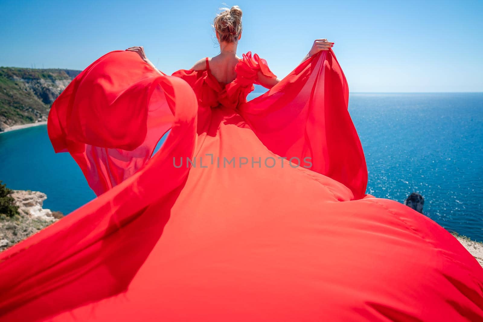 Blonde with long hair on a sunny seashore in a red flowing dress, back view, silk fabric waving in the wind. Against the backdrop of the blue sky and mountains on the seashore. by Matiunina