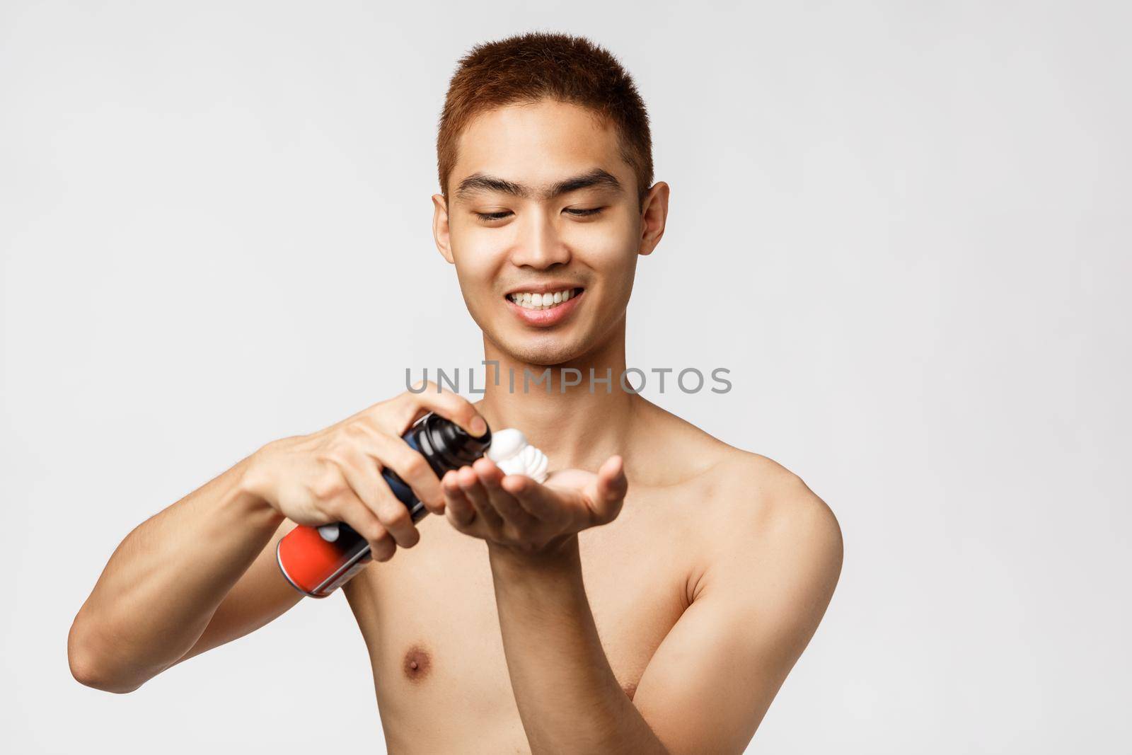 Beauty, people and hygiene concept. Portrait of handsome asian man with naked torso standing in bathroom and use shaving cream, spray foam on hand, smiling pleased, white background by Benzoix