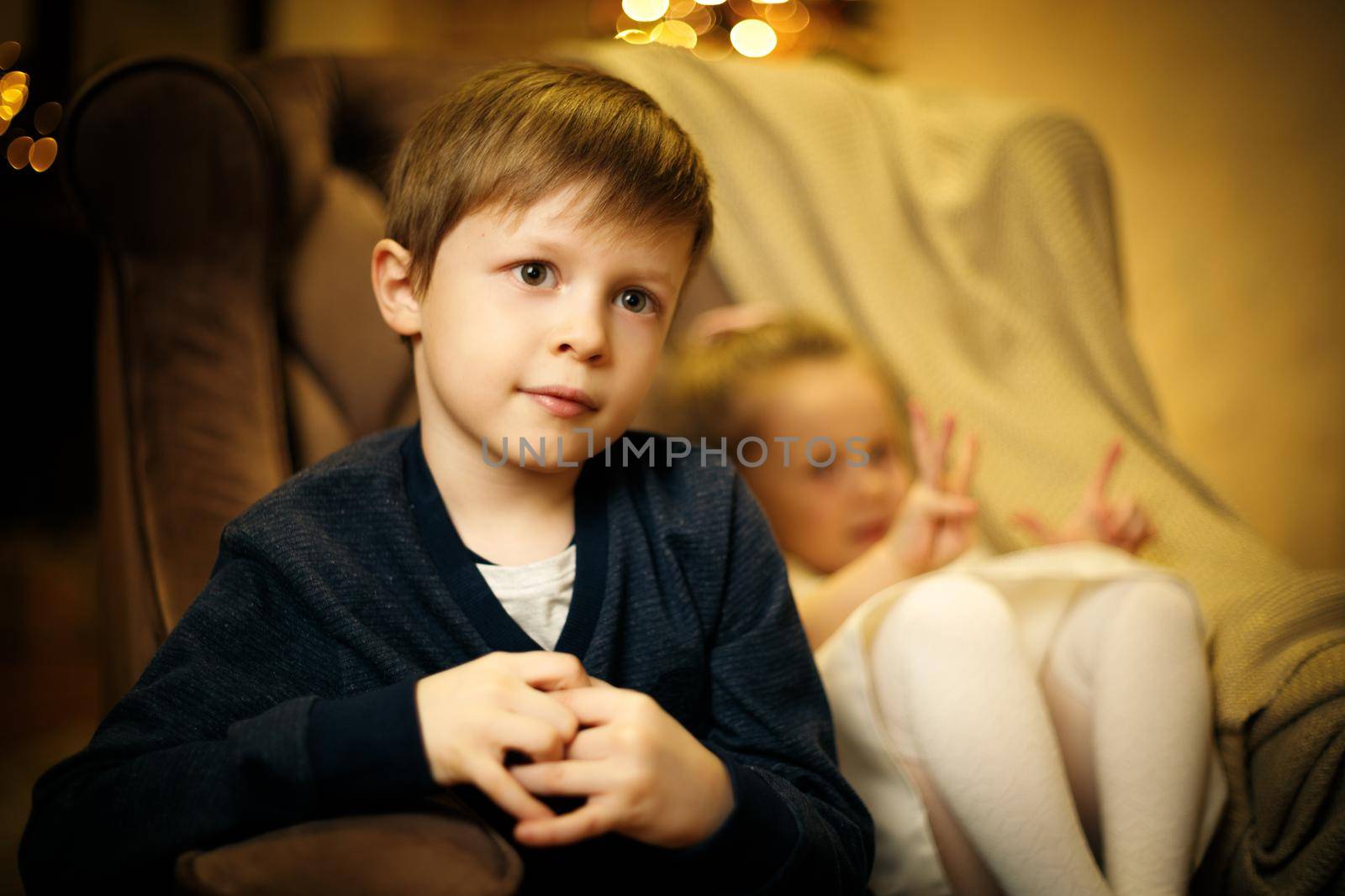 Portrait of a blond boy 5-7 years old in a chair with a little sister out of focus. Selective soft focus, film grain effect