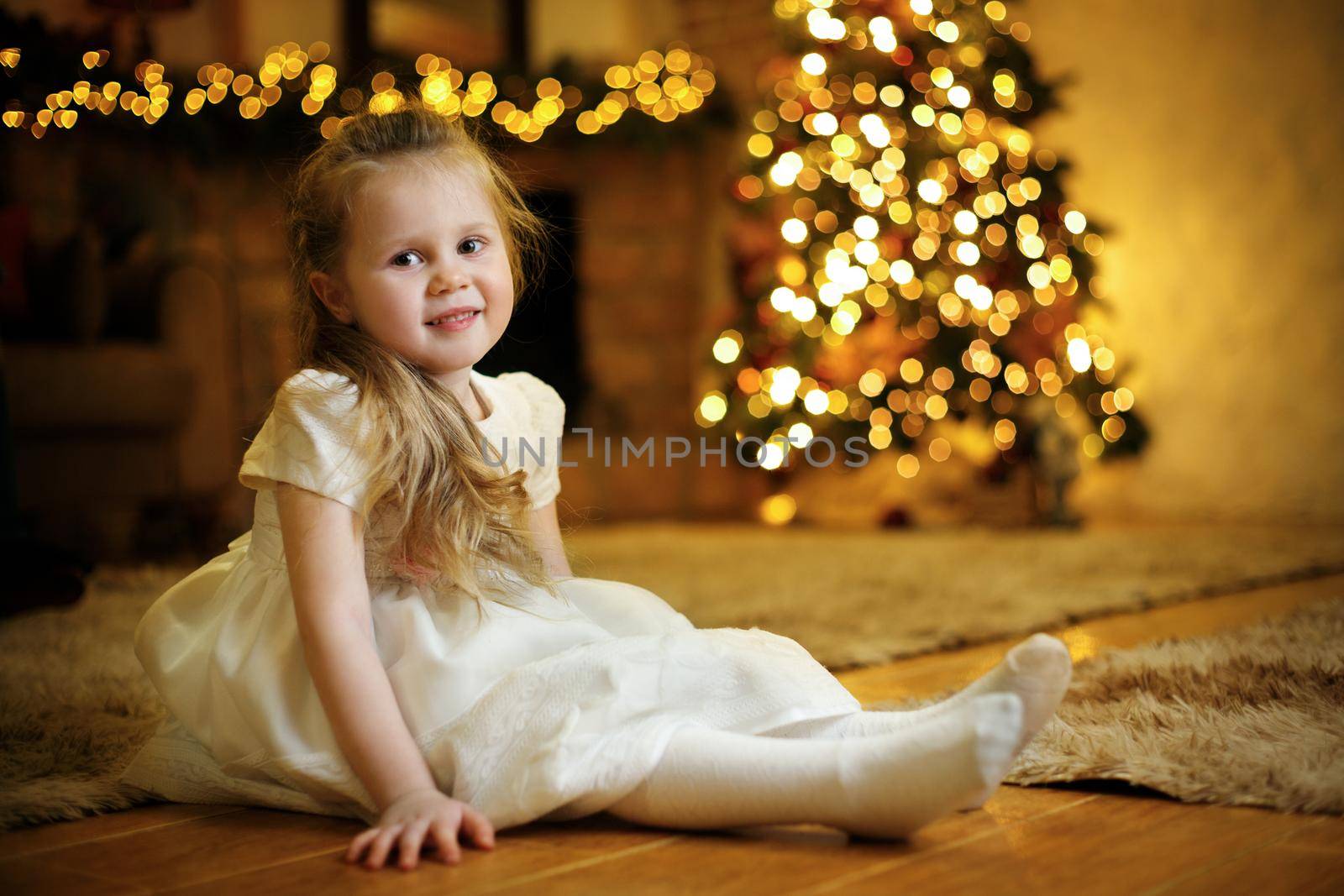 Portrait of a blond cute girl 3-5 years old sitting on the floor against the background of a festive Christmas tree. Selective soft focus, film grain effect