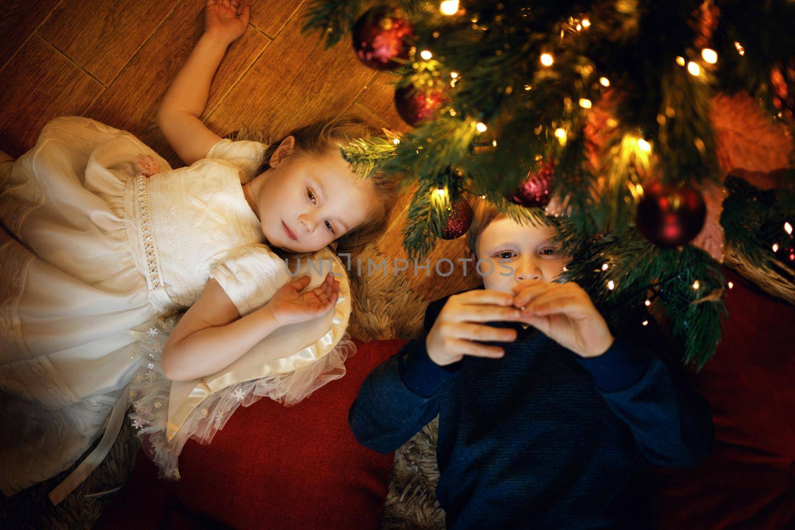 Brother and sister lies on the carpet near the New Year tree in cozy christmas interior with festive garland. Selective soft focus, film grain effect
