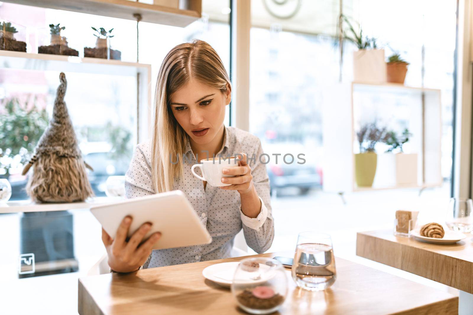 Beautiful young smiling woman drinking coffee in a cafe. She is surfing the internet on a digital tablet.