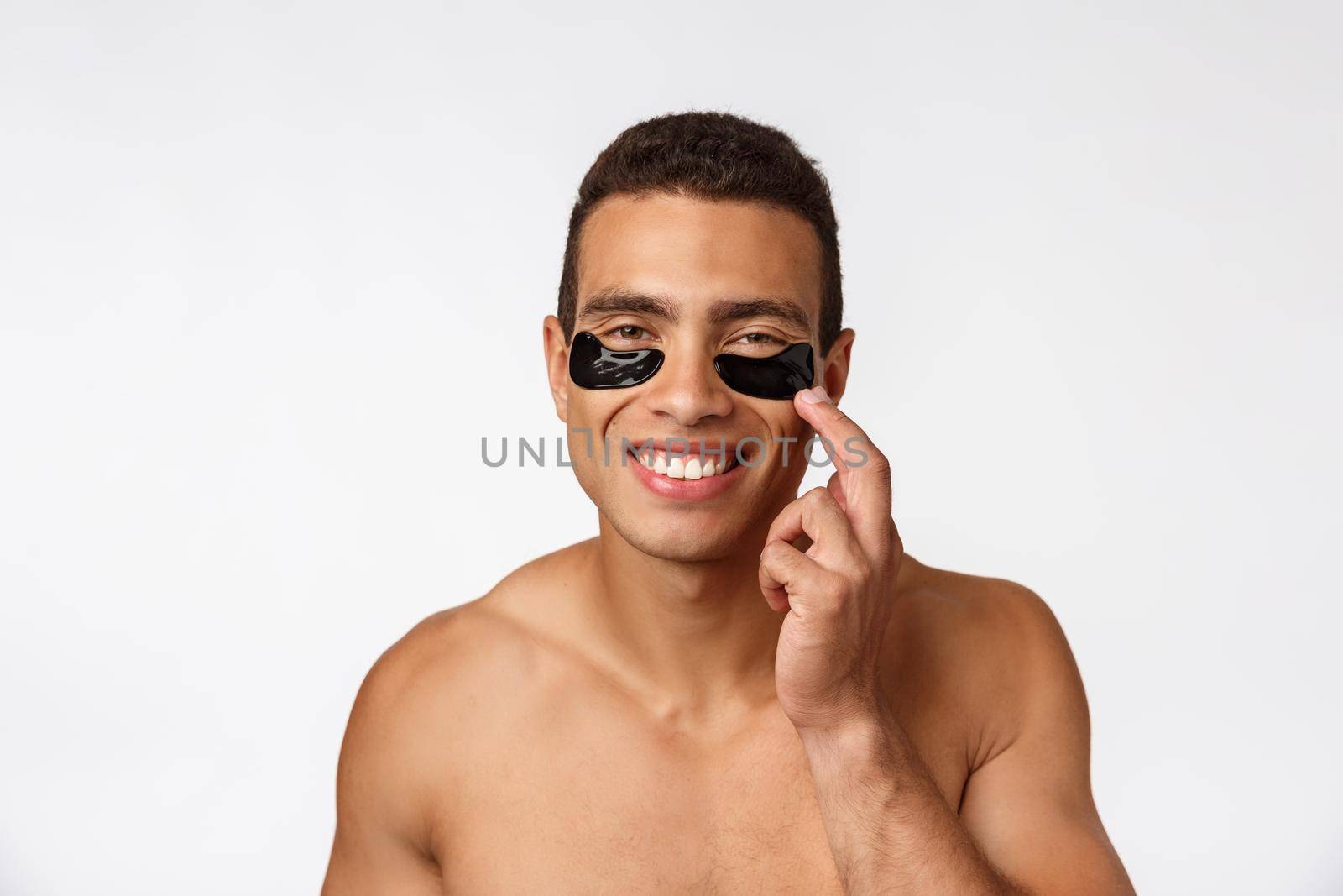 Attractive african young man with under eye black patches caring for his facial skin looking straight at camera on grey background.