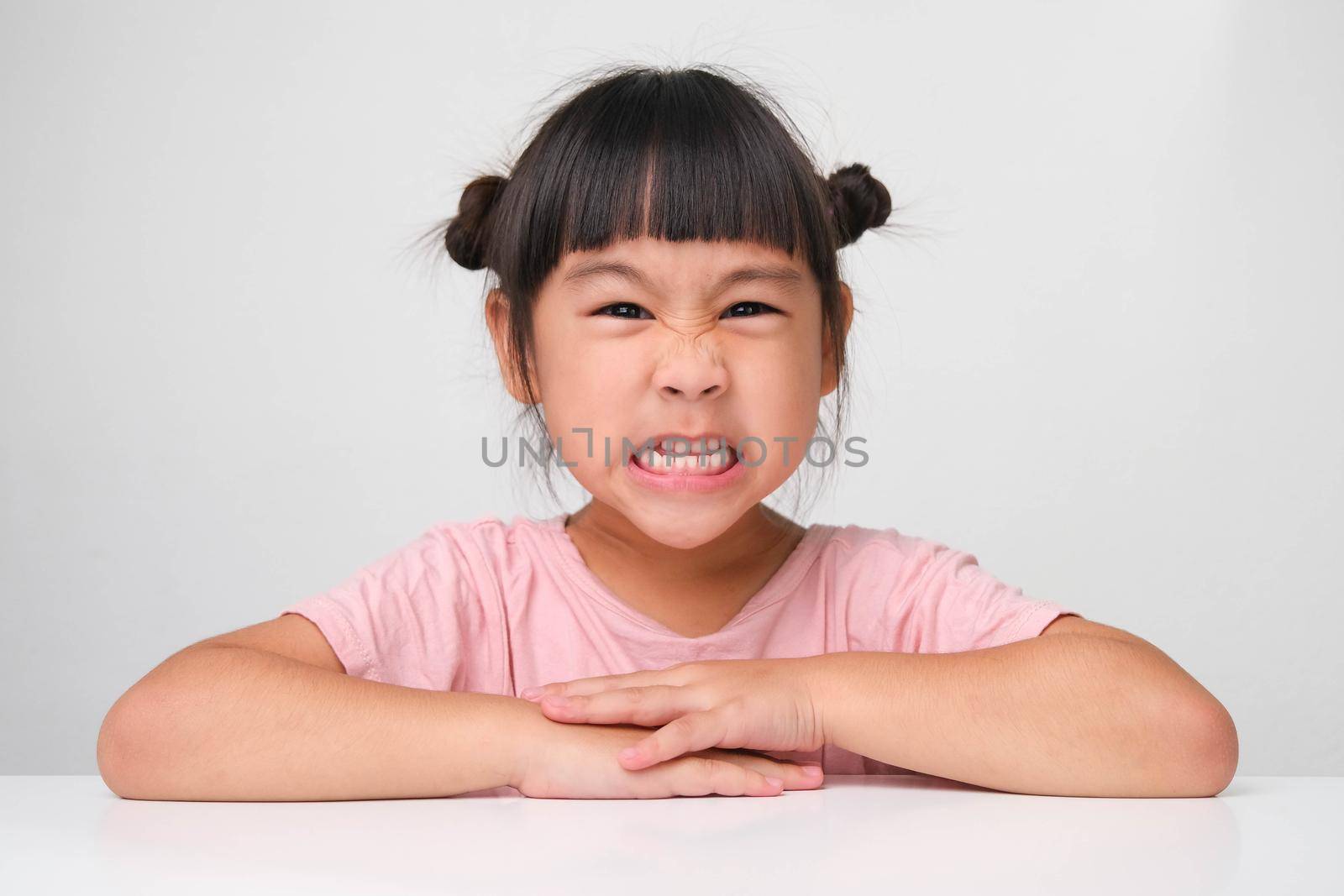 Cute dark haired little girl with angry face sitting at a table on a white background and looking at the camera.