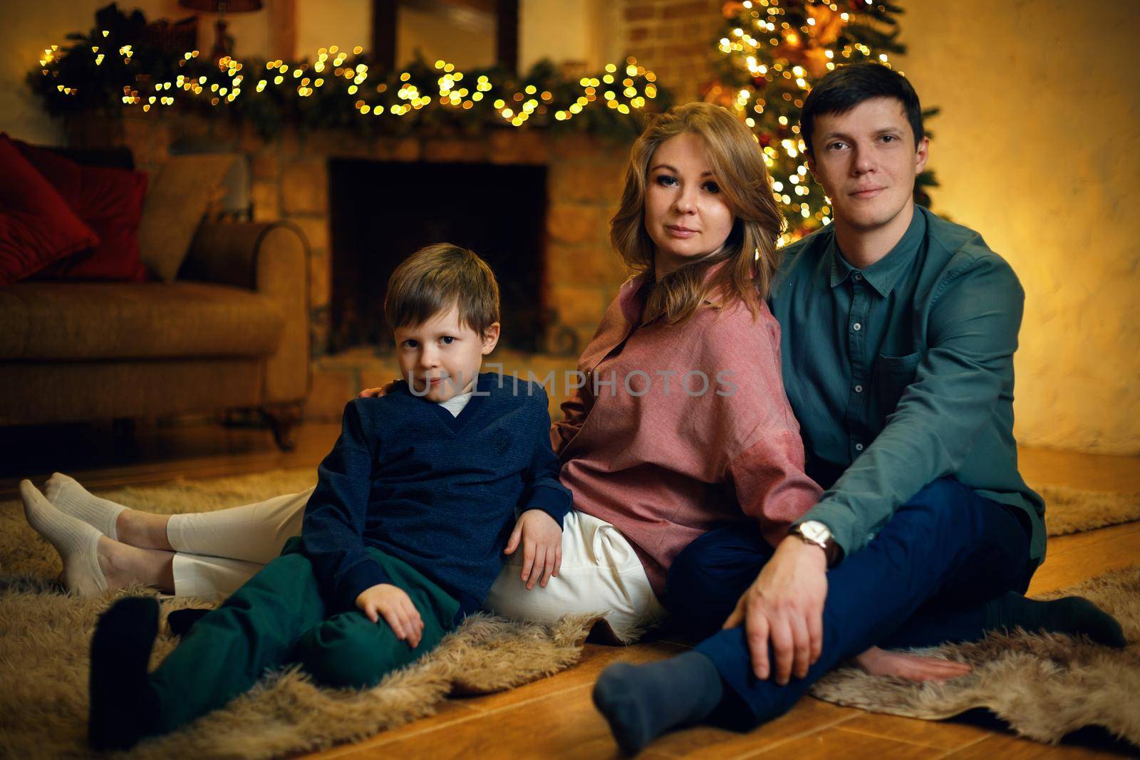 Young beautiful caucasian couple with their son posing on floor in a cozy Christmas interior with a christmas tree and garlands. Selective soft focus, film grain effect