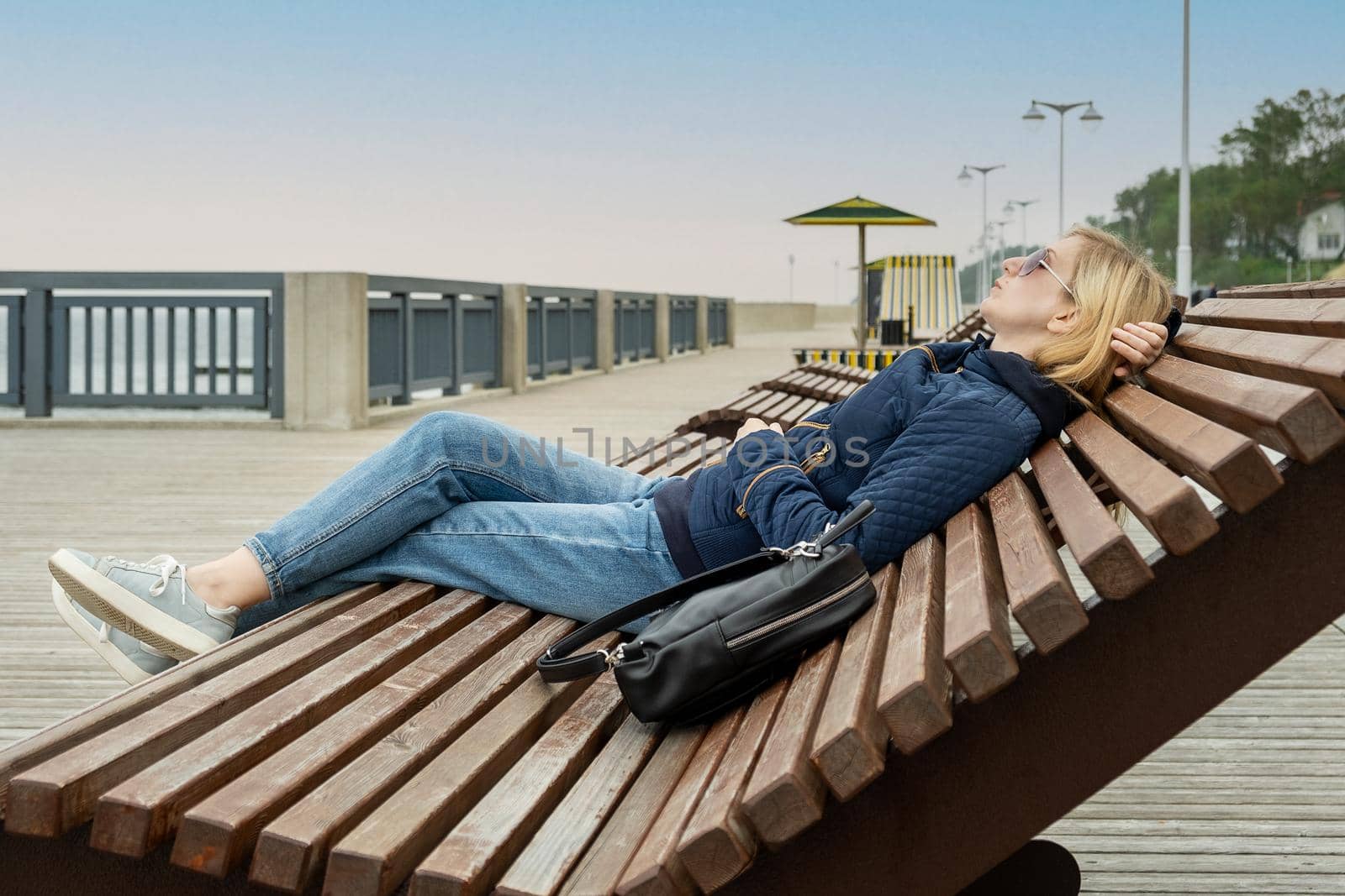 Young lonely blonde woman on an empty city embankment rests with her eyes closed on a sun lounger on spring or autumn evening. Girl misses the warmth and summer. Selective focus.