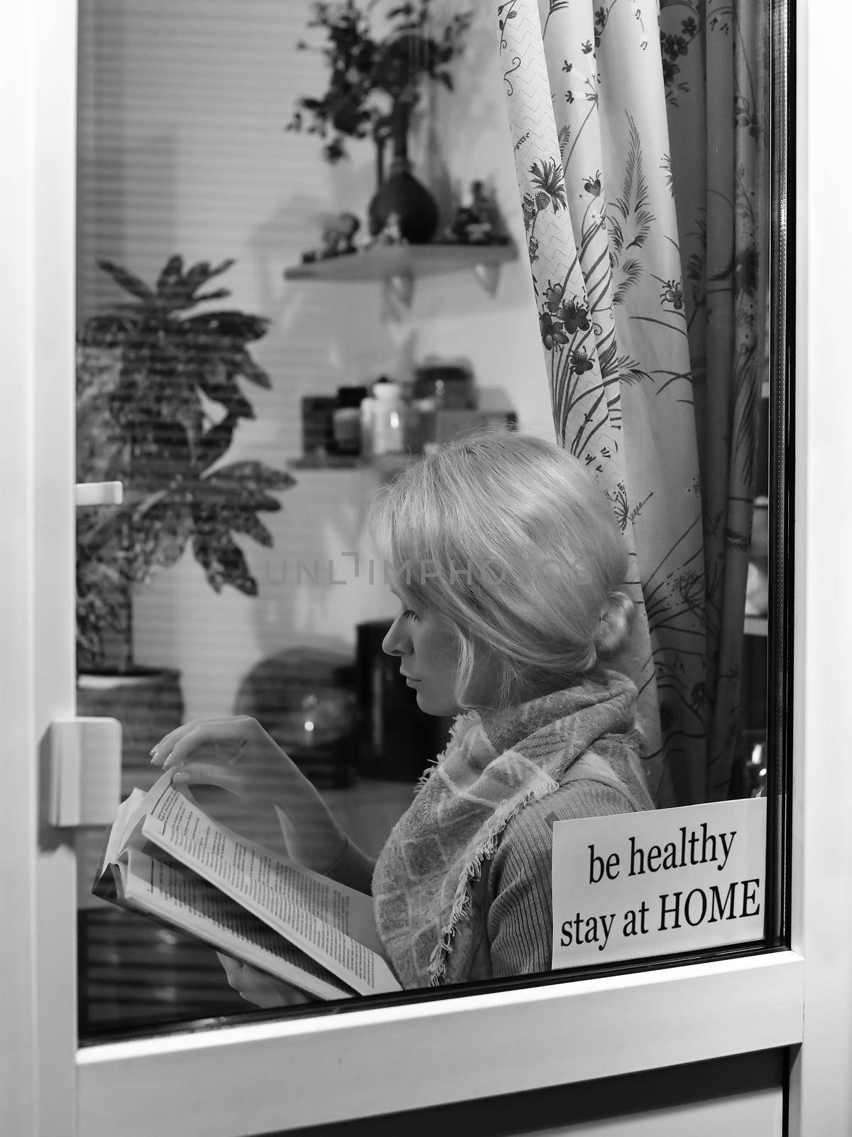 Blond woman stay home and reads book near window. Self isolating while quarantine concept. Education and rest at home by MarinaFrost