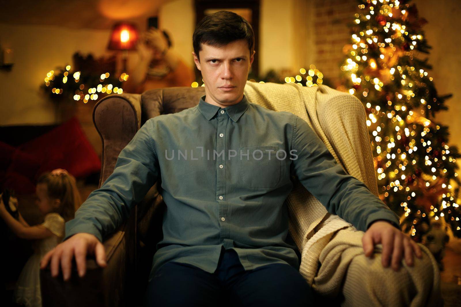 Portrait of a funny serious tired young father in an armchair, against the background of a christmas tree. Selective soft focus, film grain effect
