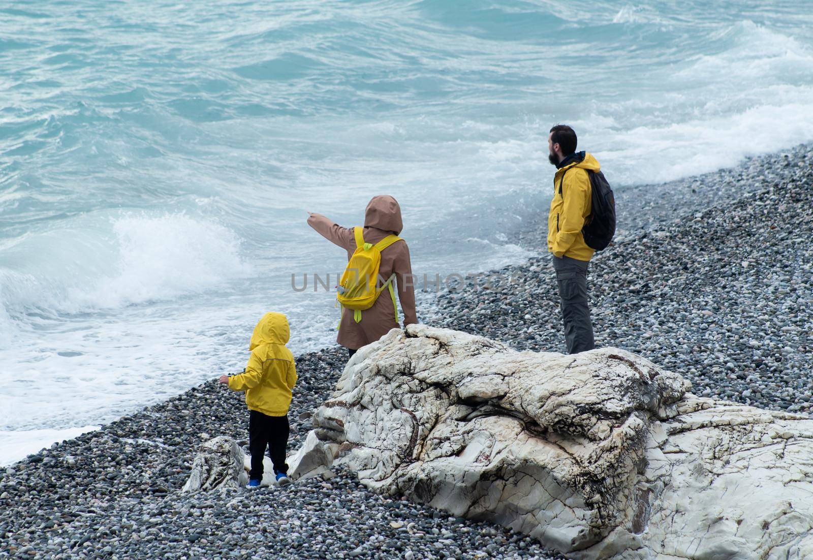 SOCHI, RUSSIA - MAY 09, 2022: a man, a woman and a child walk along a pebble beach in yellow raincoats by Rotozey