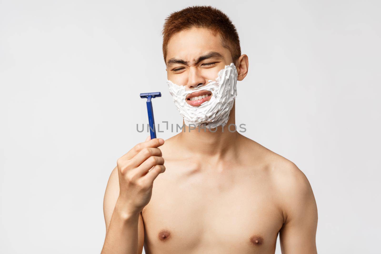 Beauty, people and hygiene concept. Portrait of disappointed, skeptical asian man grimacing, looking at razor displeased as it bad, shaving with old blades, apply facial cream to get rid of moustache by Benzoix