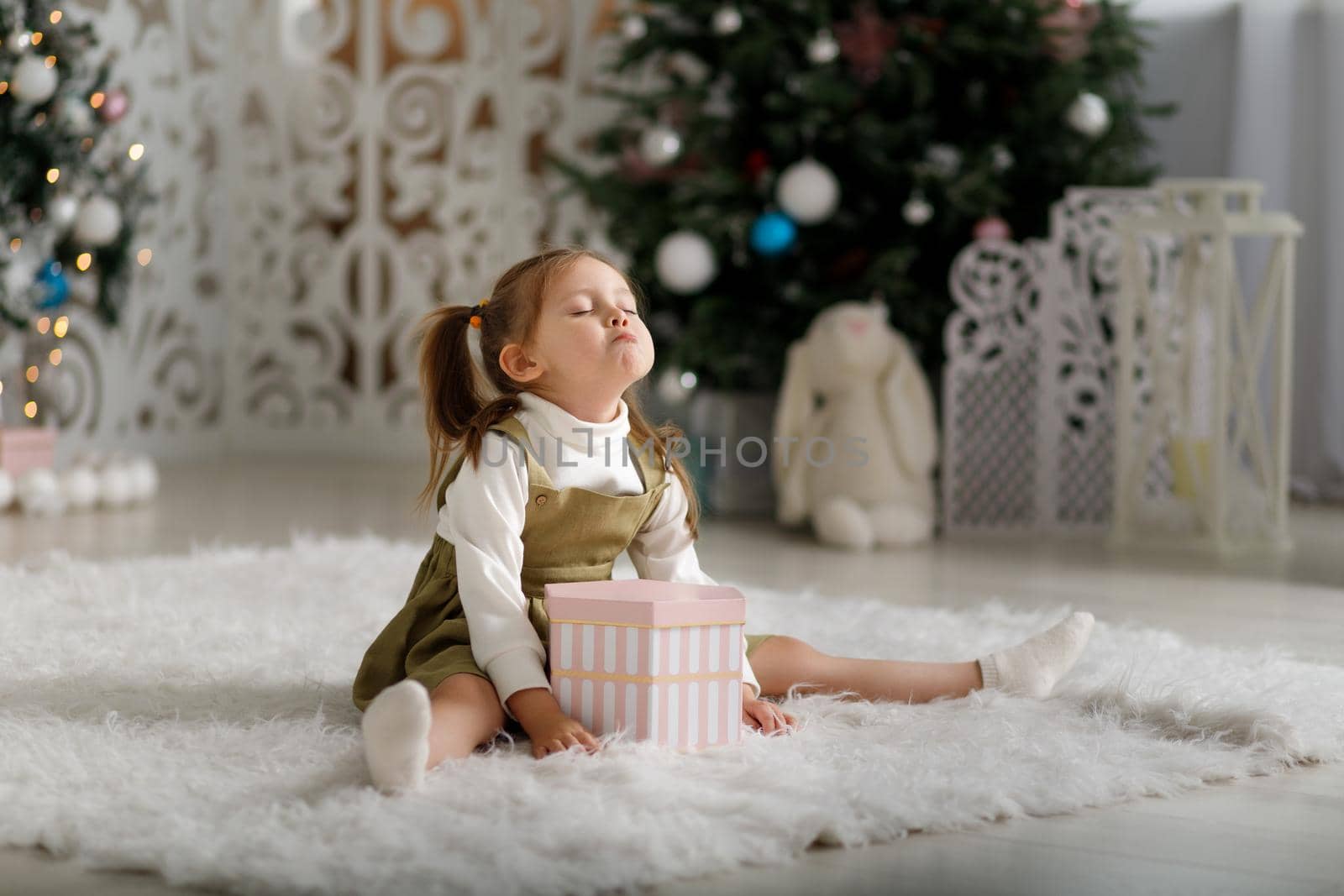Offended little girl on a white carpet with new year gift against the background of Christmas trees.