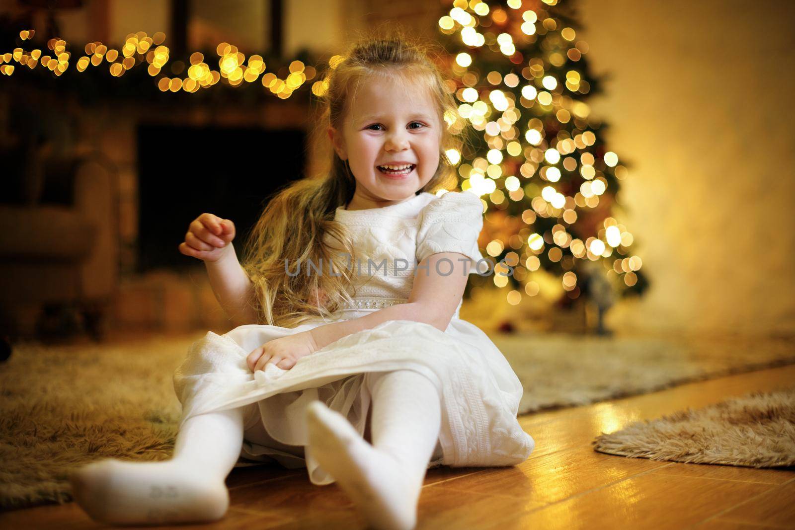 Happy little cute girl 3-5 y.o. dressed in white dress posing on floor in christmas interior with christmas tree and festive garlands. Selective soft focus, film grain effect