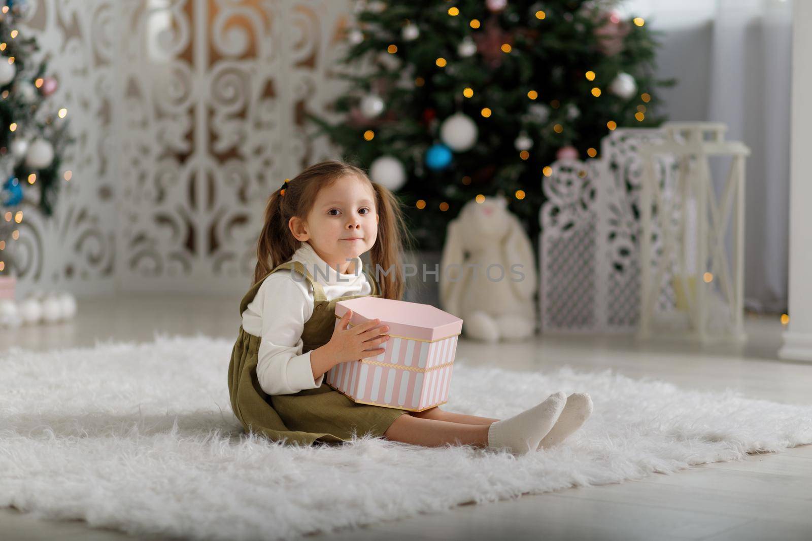 Nice pleased merry little girl holding new year gift while sitting on the carpet.