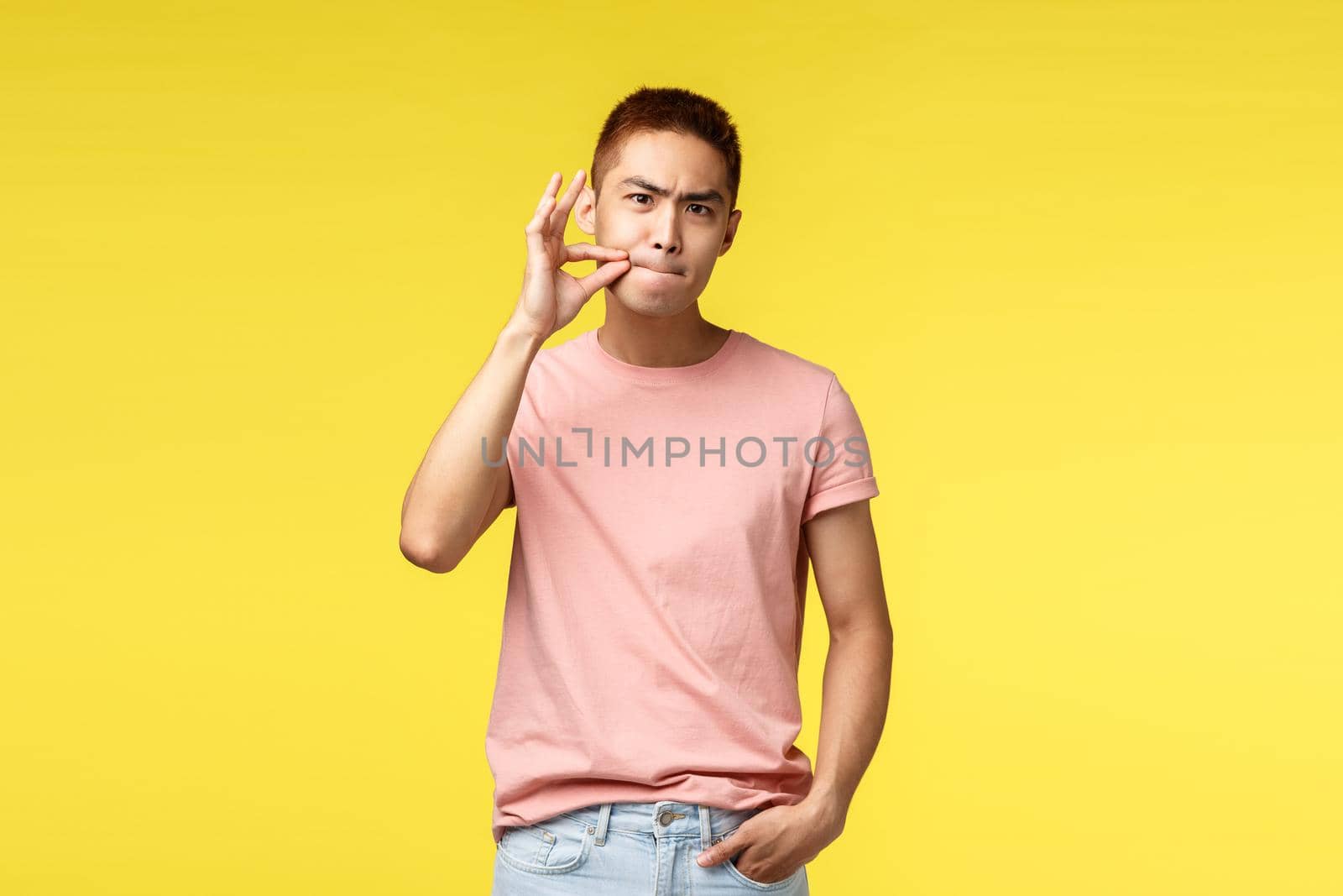 You better shut your mouth. Portrait of annoyed and serious young asian man show zip on lips, seal them as prohibit not tell anyone secret, demand silence, standing bothered yellow background.