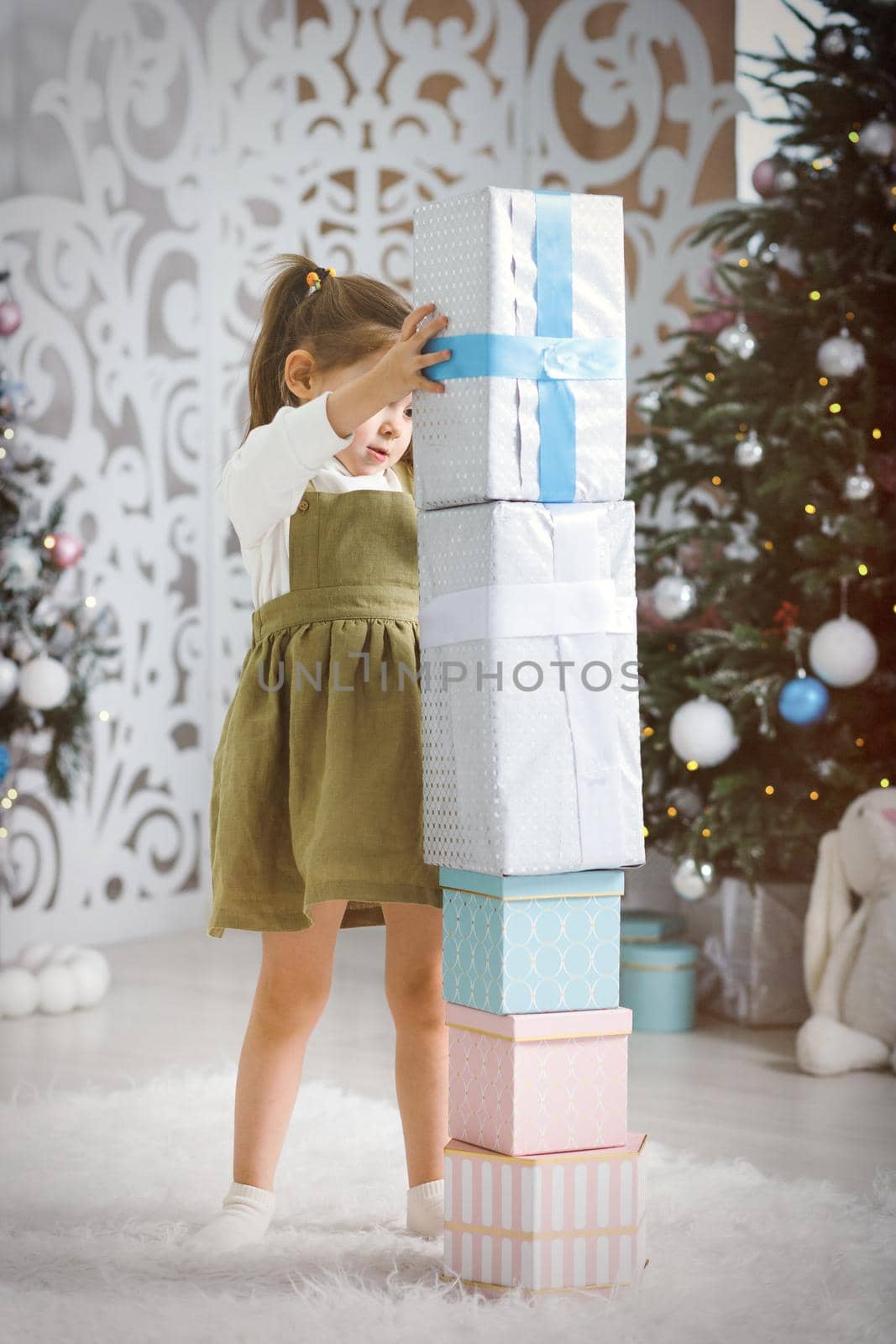 Little girl builds a tower of New Year's gifts in the living room with a Christmas tree.