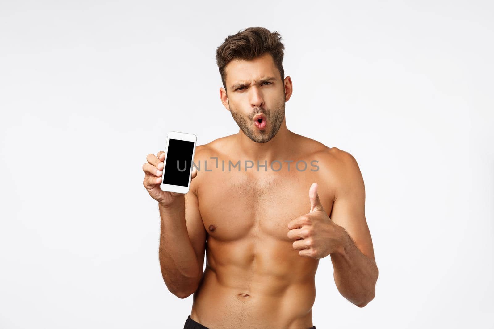 Astonished, impressed sexy young male athlete, sportsman with naked torso recommend good app, showing smartphone screen, folding lips from amazement and thumbs-up in approval, like application.