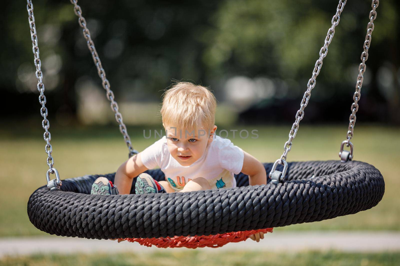 Little blonde boy playing happily on large nest swing at city playground. Round black and red seat for children's swing.