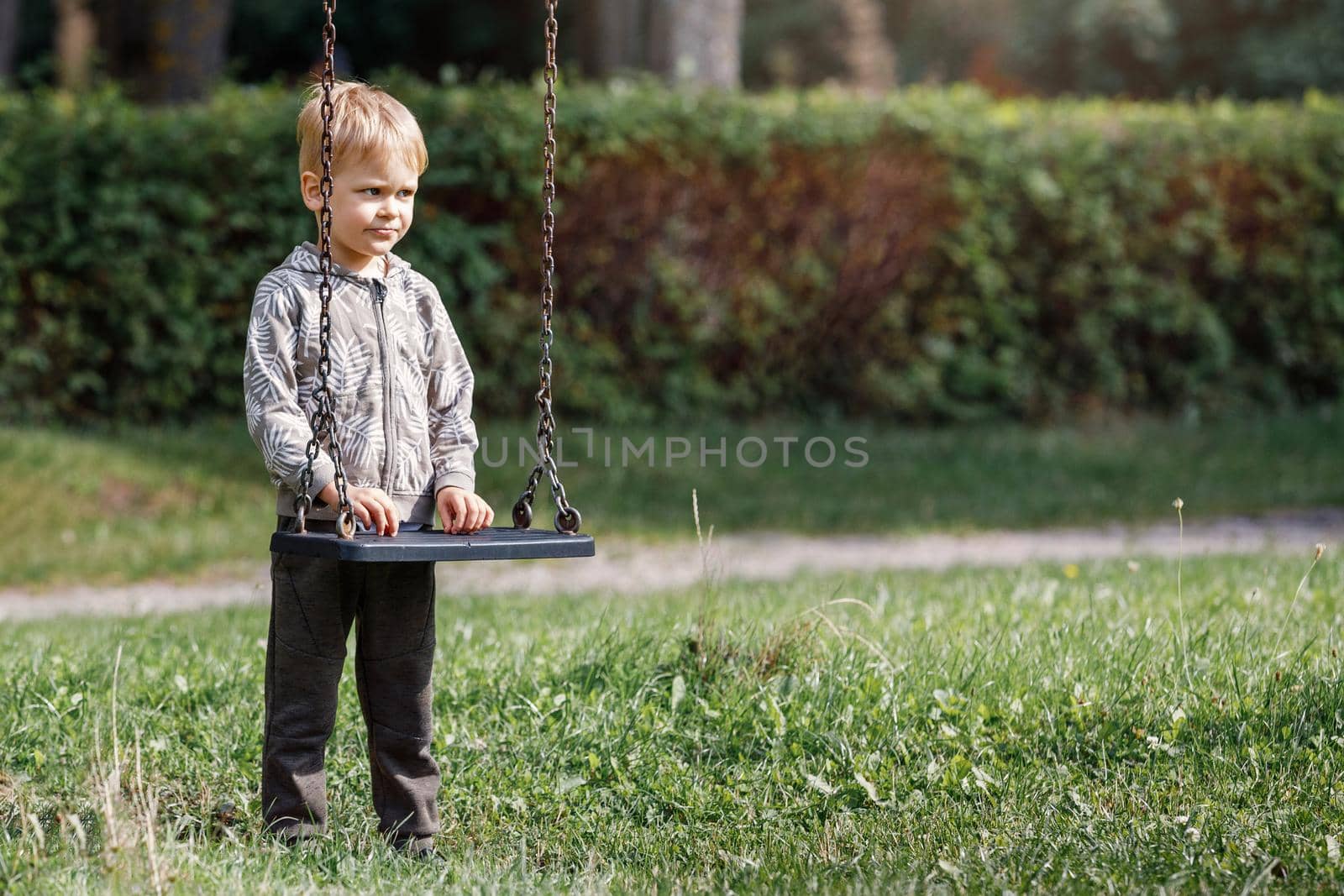 An attractive little boy stands next to a swing with chains in a city park, a child getting ready to swing. Horizontal photo by Lincikas