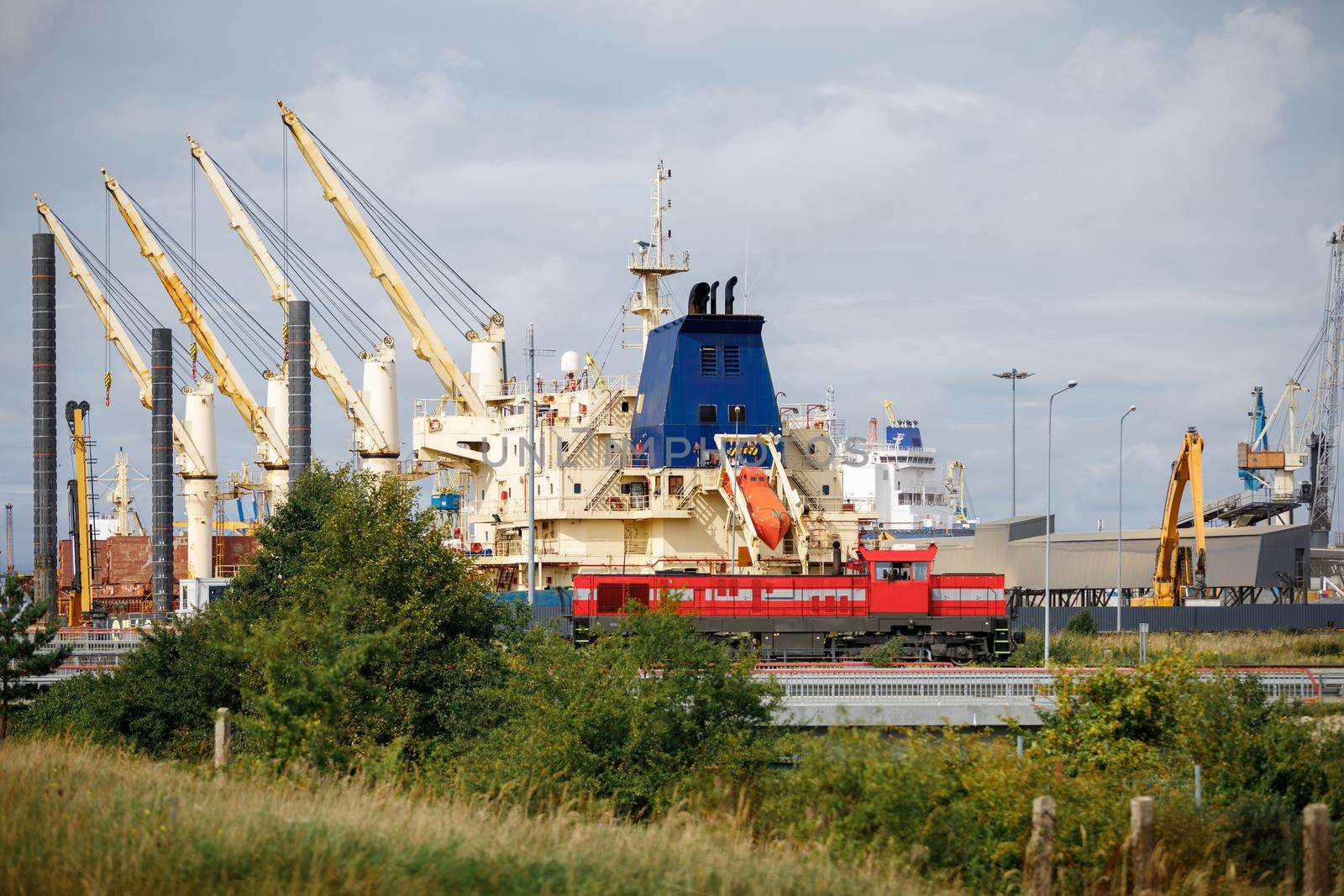 Landscape of Klaipeda port with ships, containers, ferries and cranes. In the foreground is a red steam locomotive running on a railway.