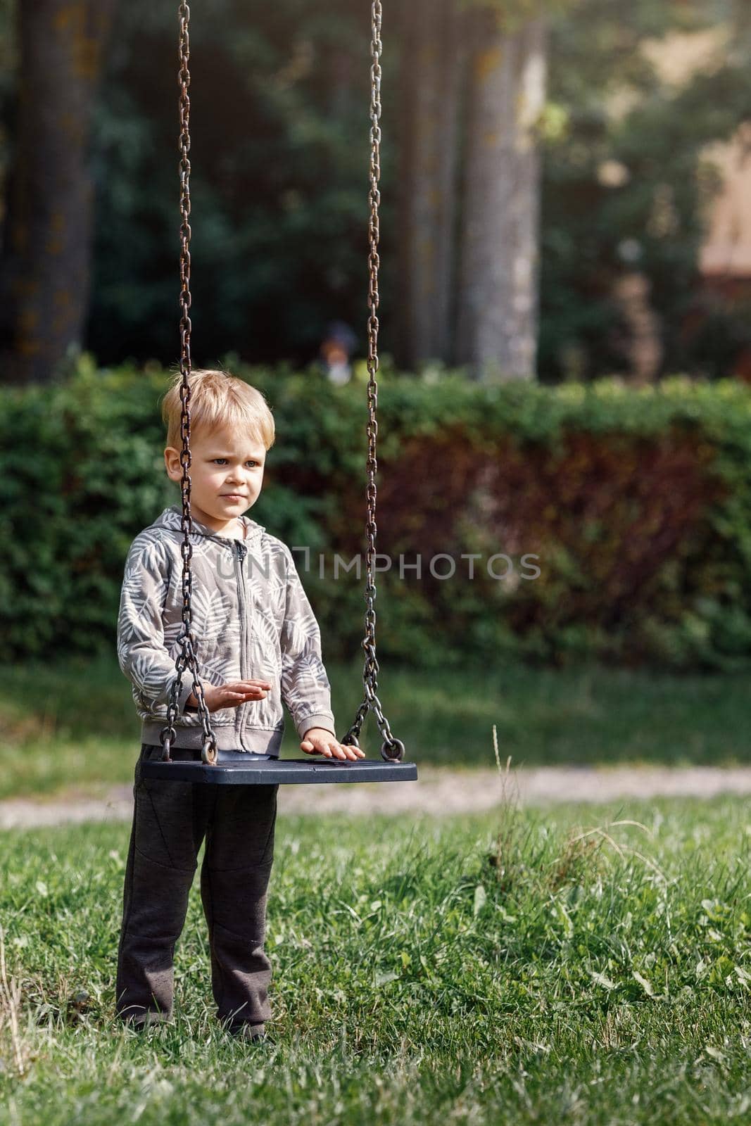 An attractive little boy stands next to a swing with chains in a city park, a child getting ready to swing. Vertical photo by Lincikas