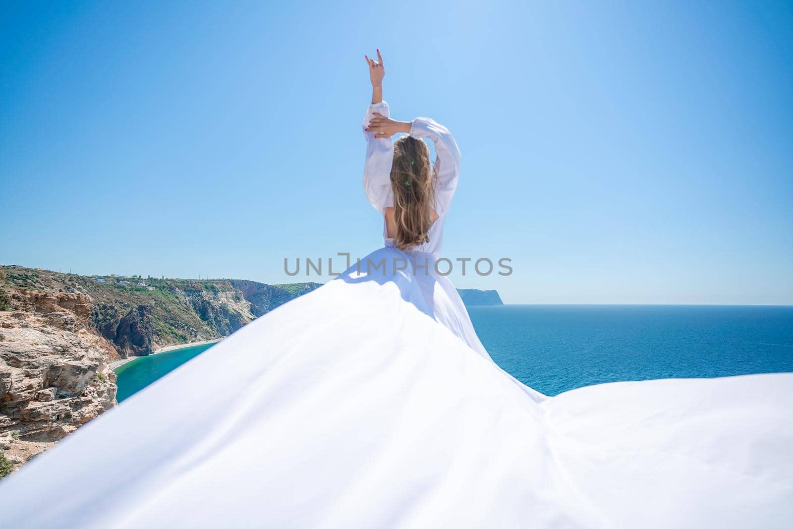 Blonde with long hair on a sunny seashore in a white flowing dress, rear view, silk fabric waving in the wind. Against the backdrop of the blue sky and mountains on the seashore