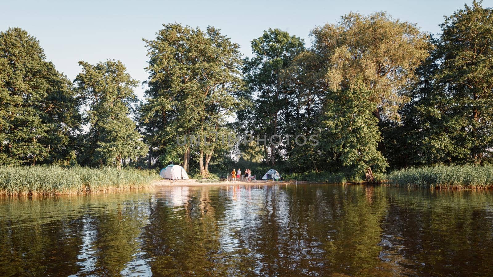 Summer camping on lake shore. Group of four young tourists standing near tent under beautiful green trees. Tourism, friendship and beauty of nature concept