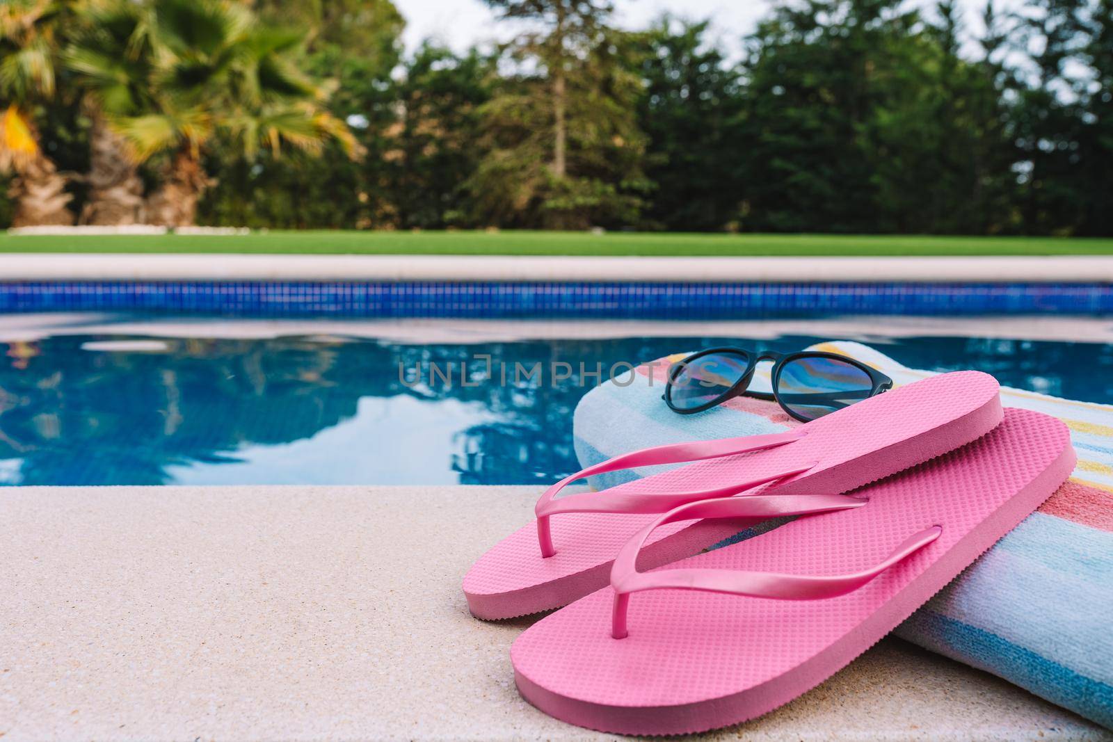 close up of summery swimming objects in front of a swimming pool. beach towel, sunglasses, swimming flip-flops. lawn garden with swimming pool in the background. by CatPhotography