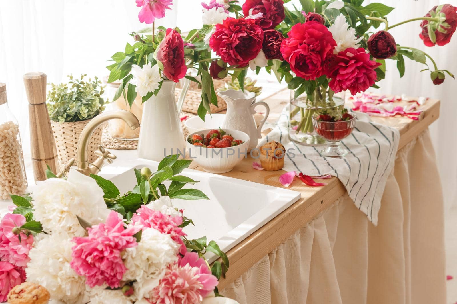 The kitchen countertop is decorated with peonies. The interior is decorated with spring flowers. Pink peonies and sweet cupcakes on a wooden countertop. Interior details.