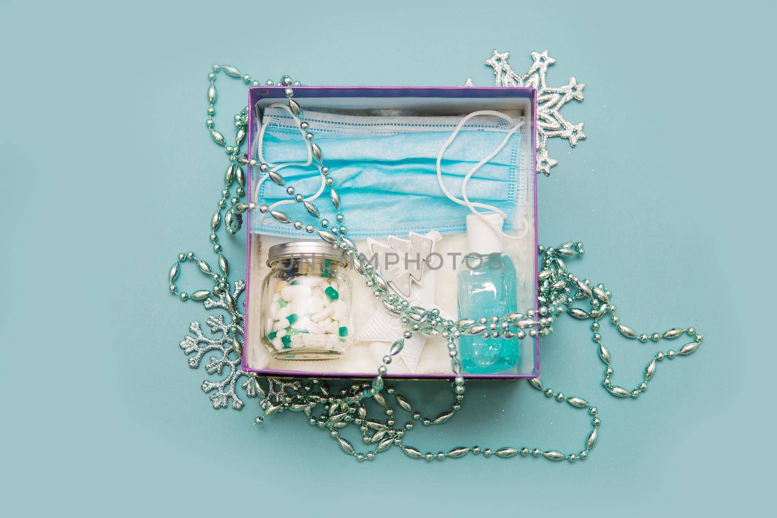 Medical mask, pills in a jar, antiseptic gel for hands and Christmas decorations in a box on a blue background. Christmas gifts in the age of coronavirus. Copy space.