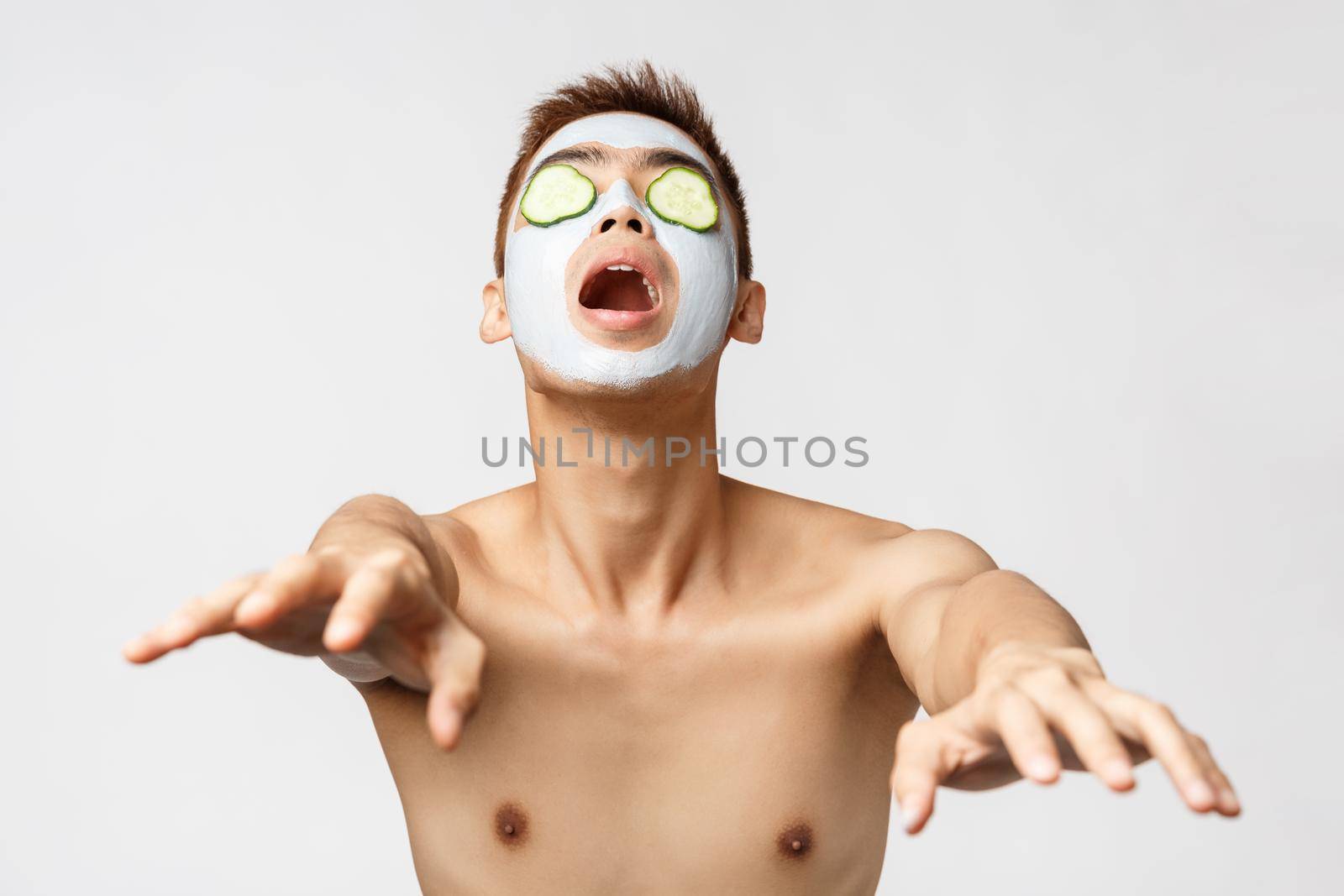 Beauty, skincare and spa concept. Portrait of funny, playful asian naked man playing around in facial mask and cucumbers on eyes, acting like zombie, reaching hands to camera, white background.